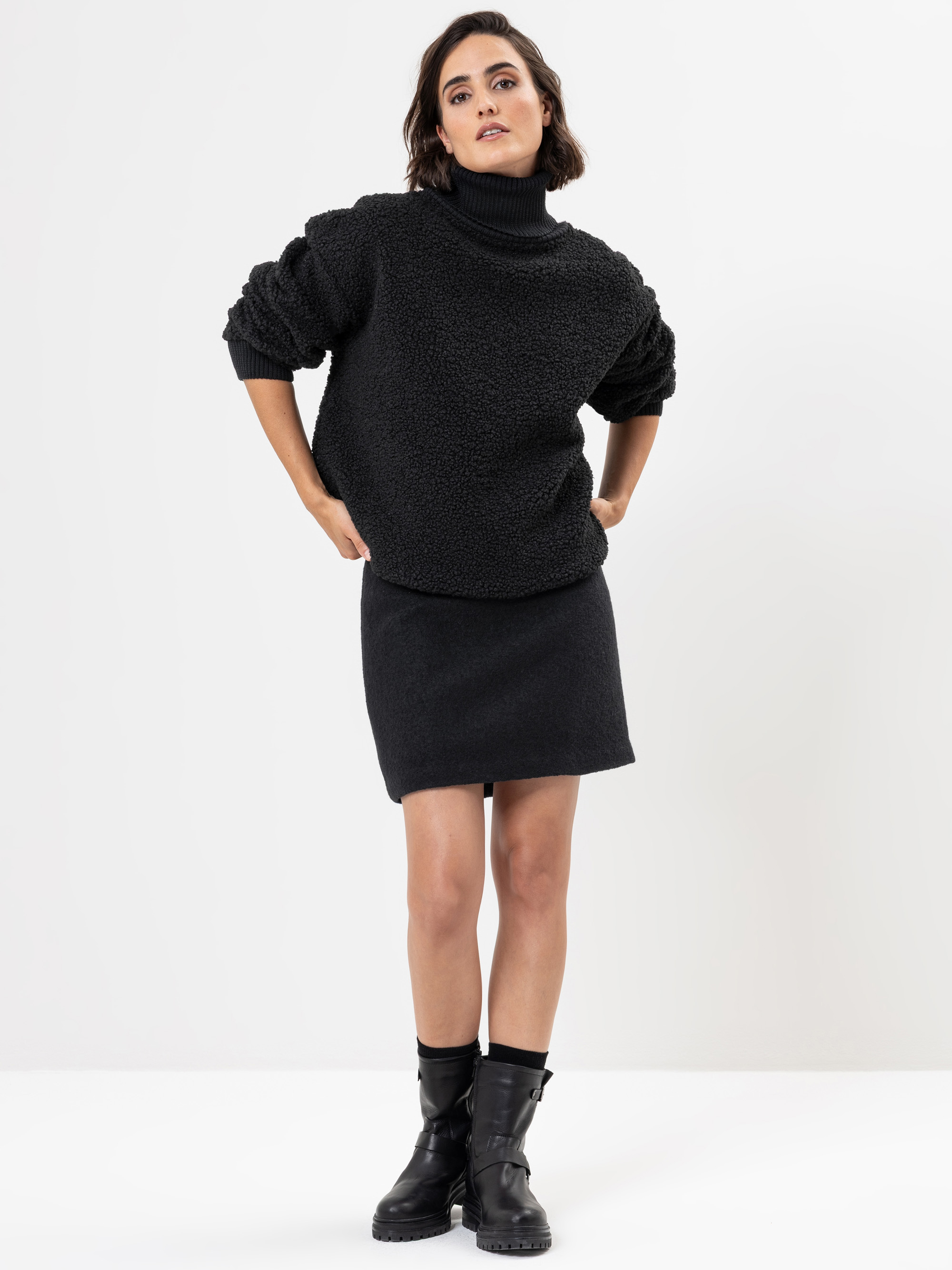 Eine Frau steht vor einem schlichten weißen Hintergrund und trägt einen schwarzen Rollkragenpullover, einen passenden schwarzen Rock und schwarze Stiefel. Ihre Hände sind in die Hüften gestemmt, und sie blickt selbstbewusst in die Kamera.