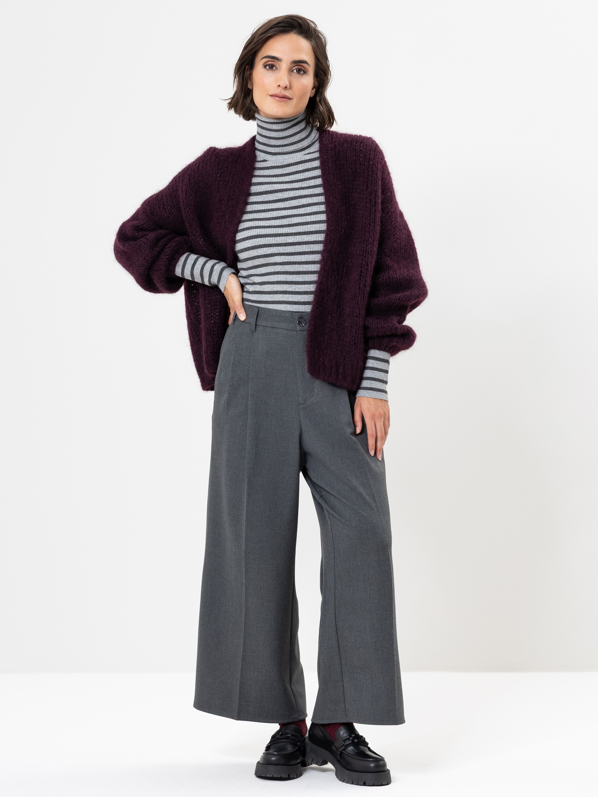 Eine Frau steht vor einem weißen Hintergrund und trägt einen gestreiften Rollkragenpullover, eine weite graue Hose, eine dicke kastanienbraune Strickjacke und schwarze Schuhe. Sie schaut mit einer Hand an der Hüfte in die Kamera.