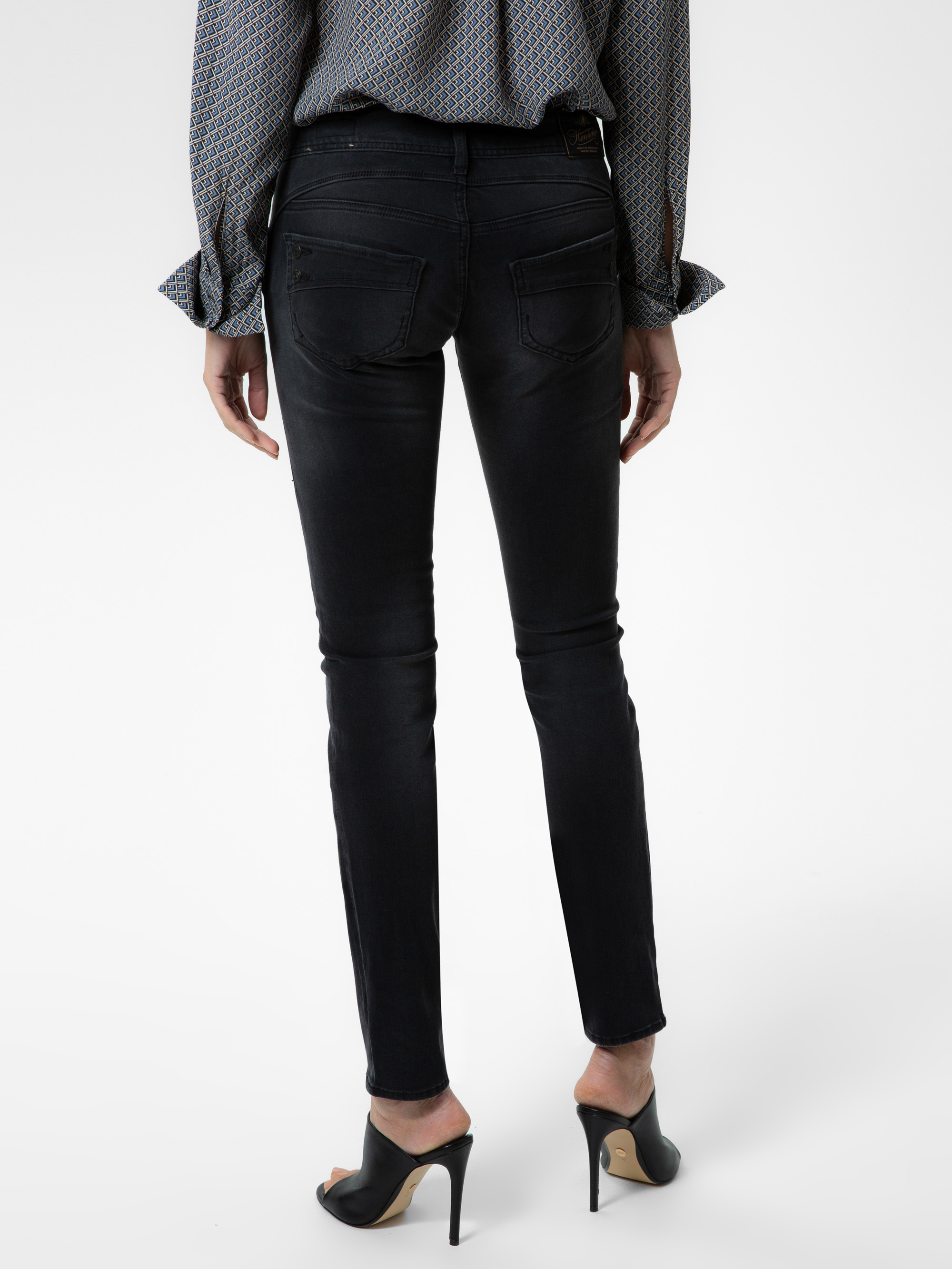 Eine Person in schwarzen Skinny-Jeans und schwarzen hochhackigen Schuhen, von hinten gesehen. Die Person trägt außerdem ein gemustertes langärmeliges Hemd mit gerüschten Manschetten.