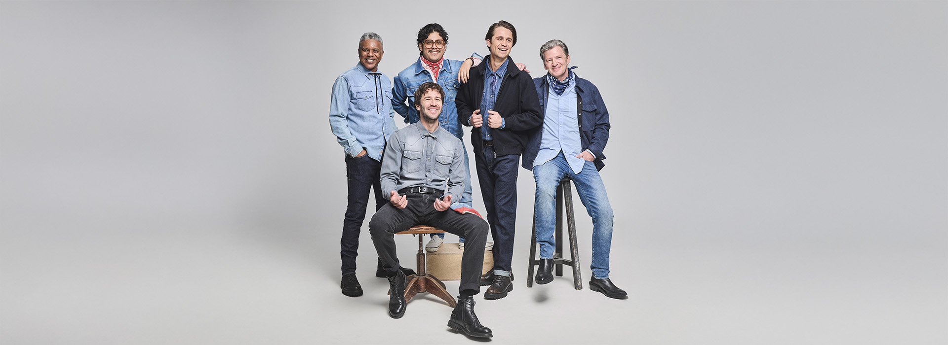 Fünf lächelnde Männer in lässigen blauen Jeans und Hemden posieren zusammen in einem Studio mit hellgrauem Hintergrund; einer sitzt auf einem Stuhl, die anderen stehen oder sitzen auf Hockern um ihn herum.