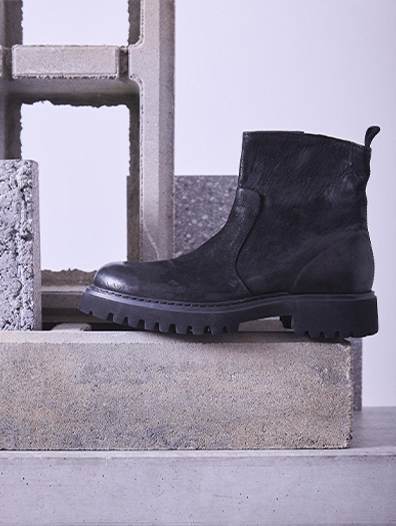 Eine schwarze Lederstiefelette mit einer robusten Sohle steht auf Betonblöcken, vor einem Hintergrund aus gestapelten Zementziegeln und einem neutralen Hintergrund.