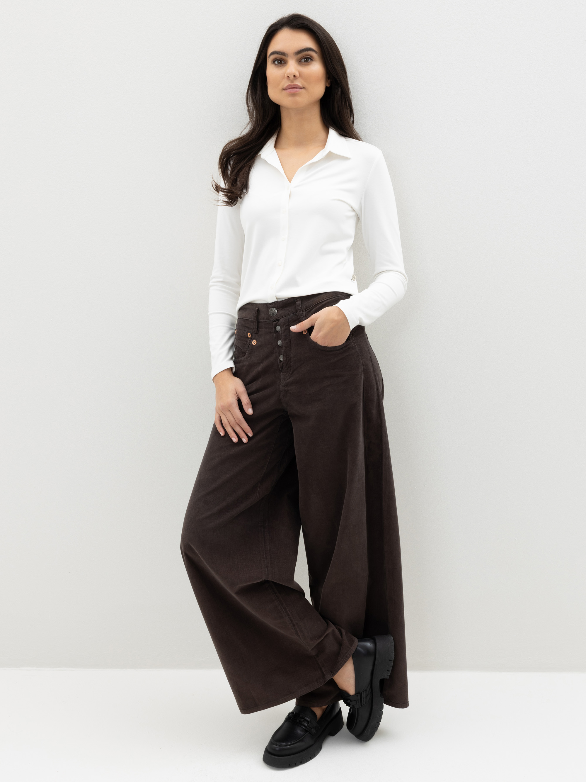 Eine Frau steht an einer weißen Wand. Sie trägt eine weiße, langärmelige Bluse, die in eine braune Hose mit hohem Bund und weitem Bein gesteckt ist, und schwarze Schuhe. Sie hat langes dunkles Haar, eine Hand in der Tasche und blickt selbstbewusst nac