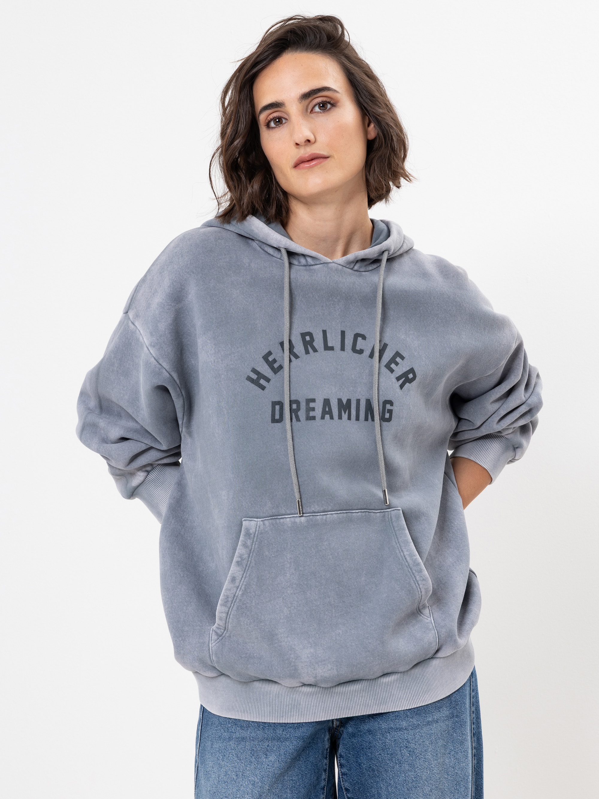 Eine Frau, die einen lockeren, hellgrauen Kapuzenpullover mit der Aufschrift HERZLICHER DREAMING trägt, steht mit den Händen in den Taschen und schaut in die Kamera vor einem schlichten weißen Hintergrund.