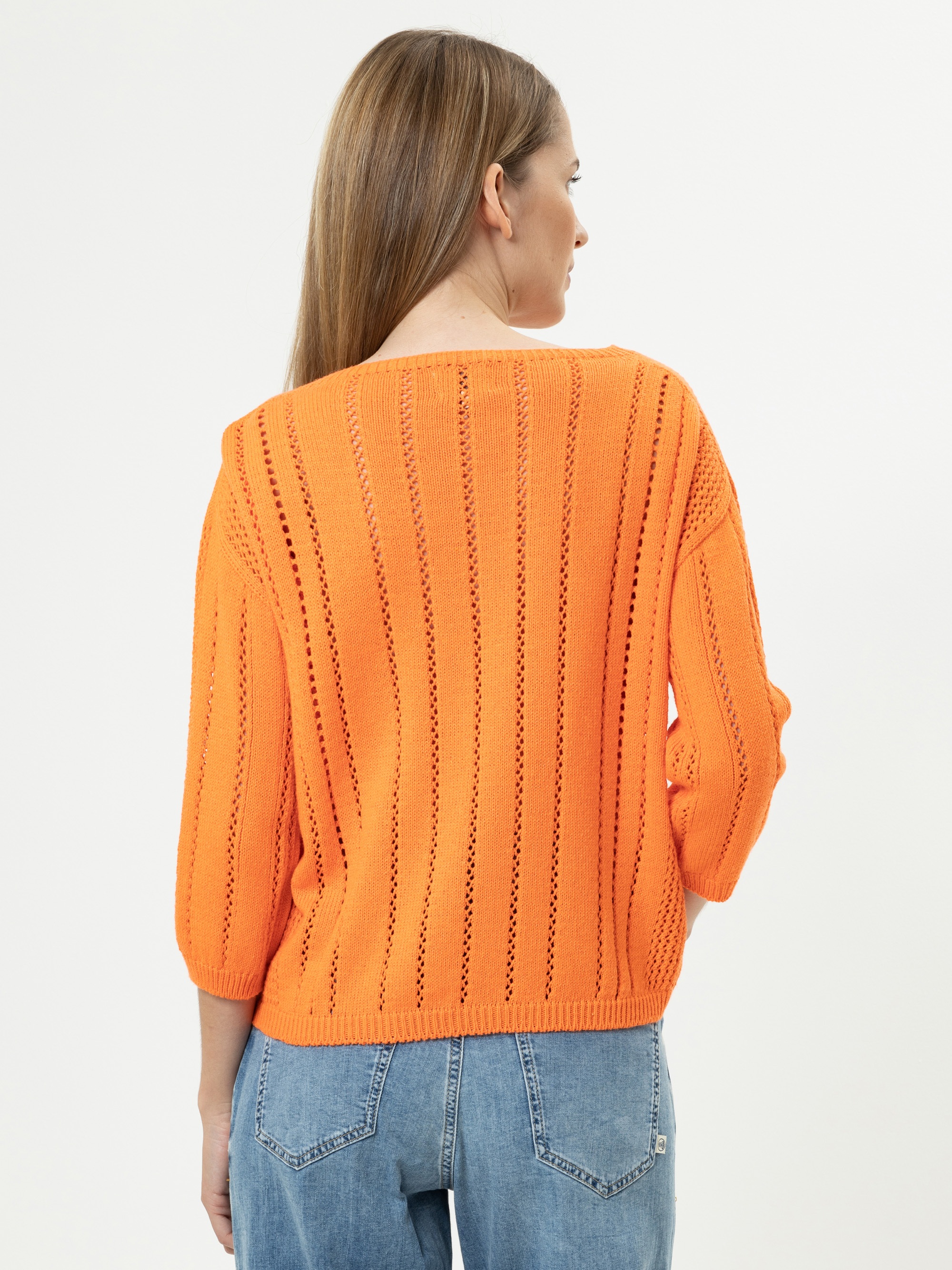 Eine Frau mit langen hellbraunen Haaren, die einen leuchtend orangefarbenen Strickpullover mit vertikalen durchbrochenen Mustern und blaue Jeans trägt, steht mit dem Gesicht zur Kamera vor einem schlichten weißen Hintergrund.