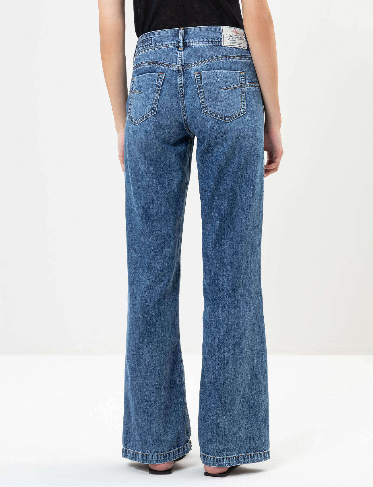 Eine Person, die eine blaue Jeans mit hoher Taille und weitem Bein und ein schwarzes Oberteil trägt, dargestellt von hinten vor einem schlichten weißen Hintergrund. Die Jeans hat Gesäßtaschen und ein Etikett am Bund.