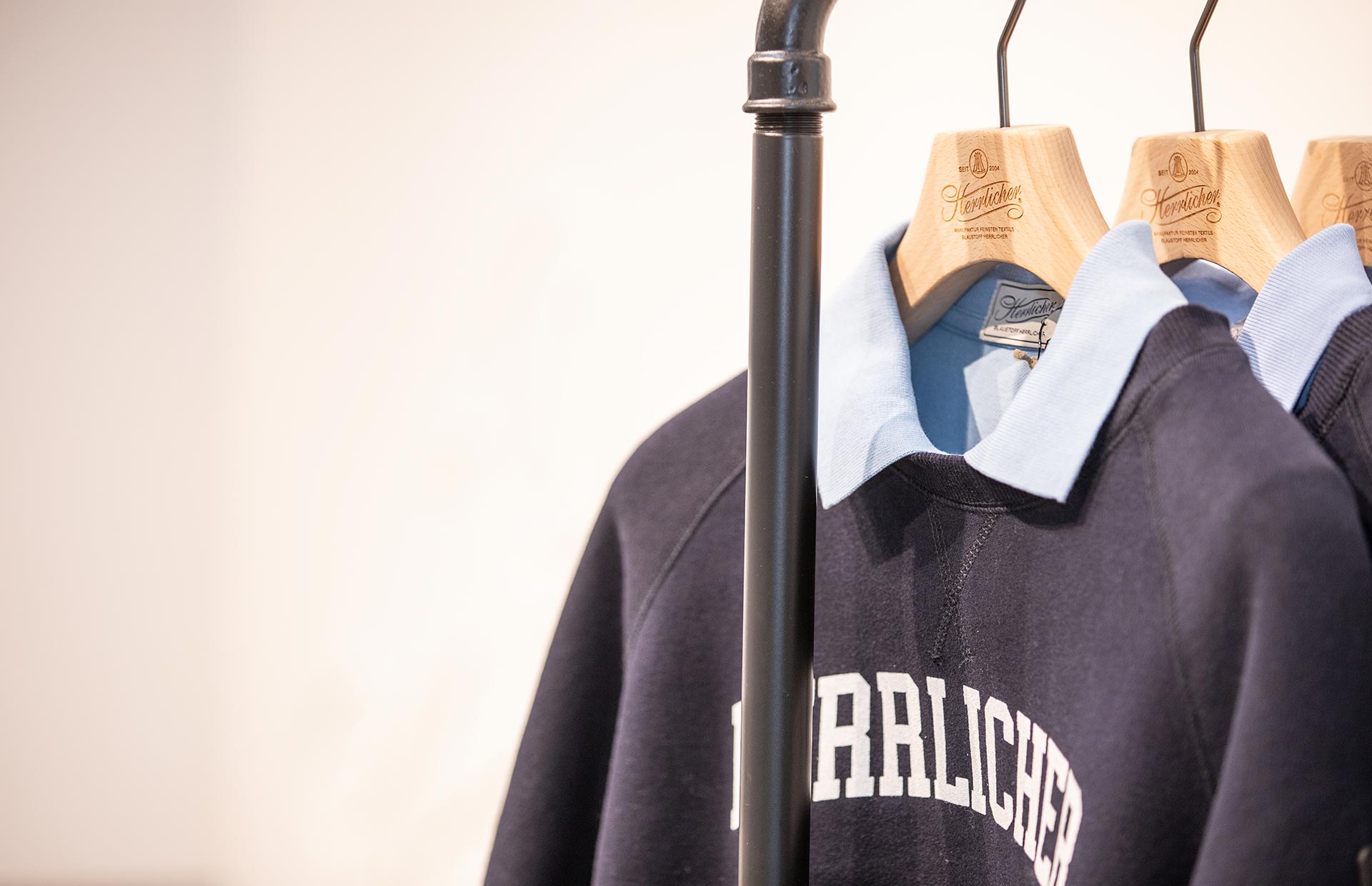 Drei marineblaue Sweatshirts mit hellblauen Kragen hängen auf Holzbügeln an einem schwarzen Kleiderständer. Das sichtbare Sweatshirt hat einen weißen Schriftzug auf der Brust, auf dem teilweise RRLICHE steht.