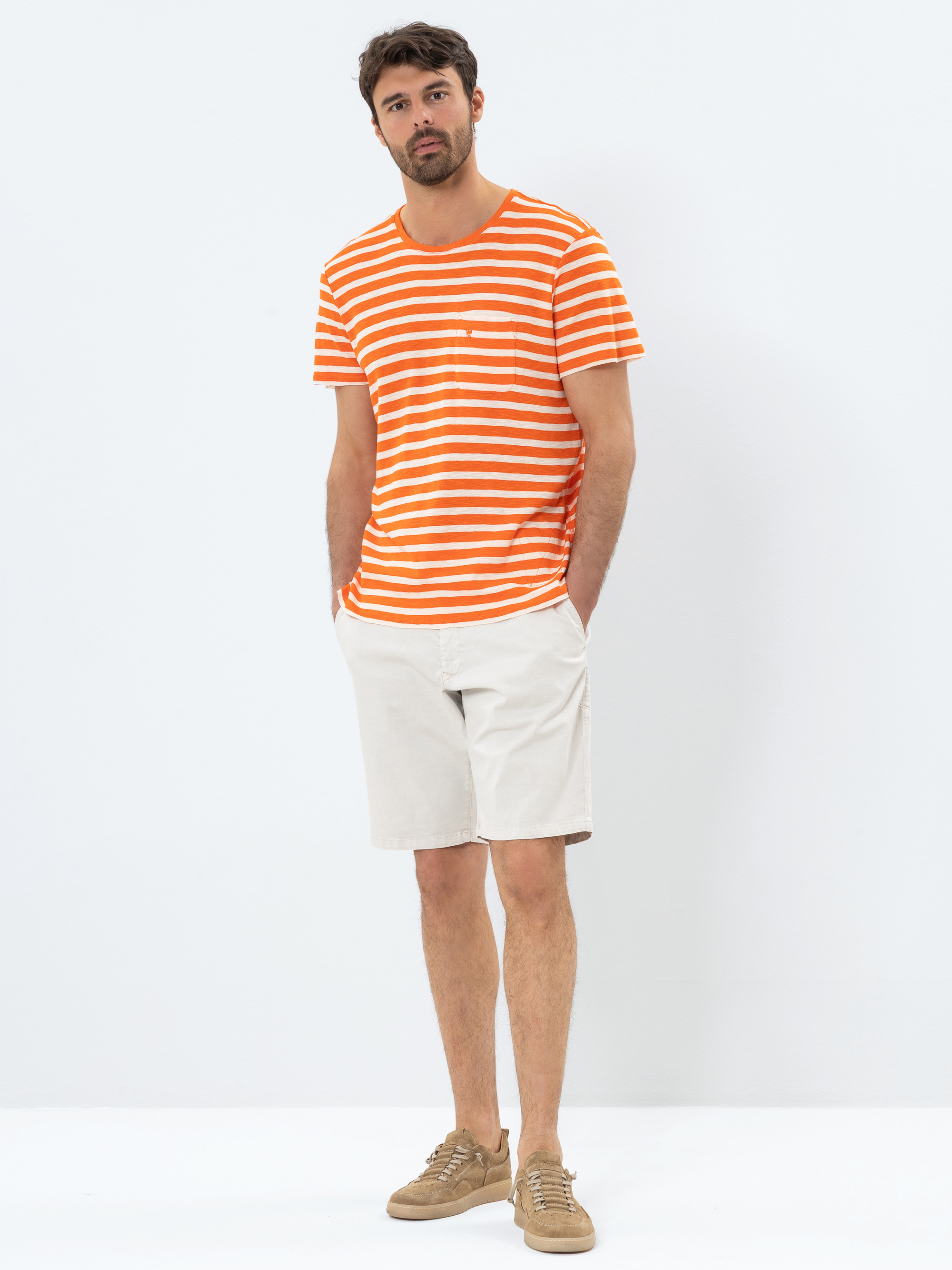 Ein Mann steht vor einem schlichten weißen Hintergrund, trägt ein orange-weiß gestreiftes T-Shirt, hellbeige Shorts und hellbraune Turnschuhe und hat die Hände in den Taschen.