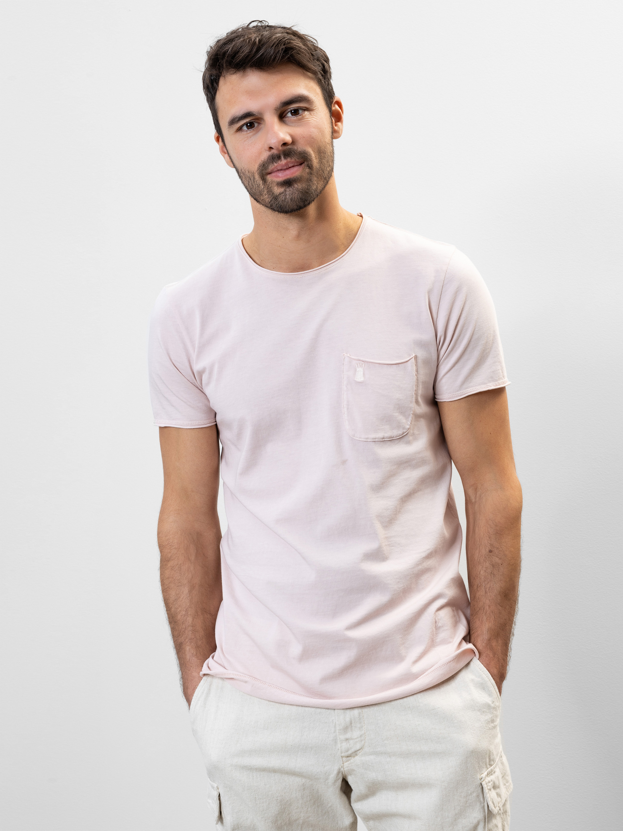 Ein Mann mit kurzen dunklen Haaren und einem Bart trägt ein hellrosa T-Shirt mit einer Brusttasche und eine helle Hose. Er steht vor einem schlichten weißen Hintergrund, die Hände in den Taschen, und schaut mit neutralem Blick in die Kamera.