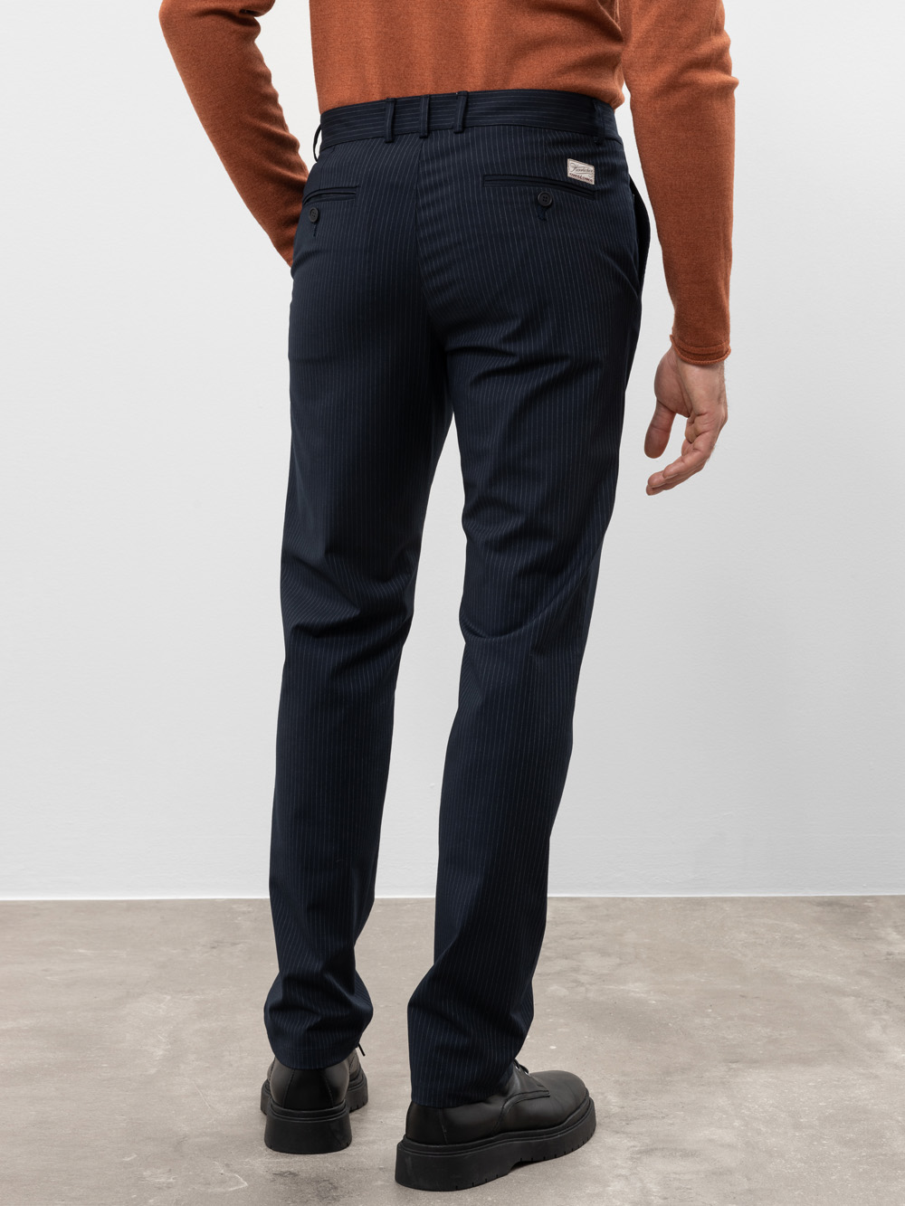 Eine Person, die einen rostfarbenen Pullover und eine dunkle Nadelstreifenhose trägt, steht auf einem grauen Boden und schaut von der Kamera weg. Die Arme sind entspannt an der Seite und die Person trägt schwarze Schuhe.