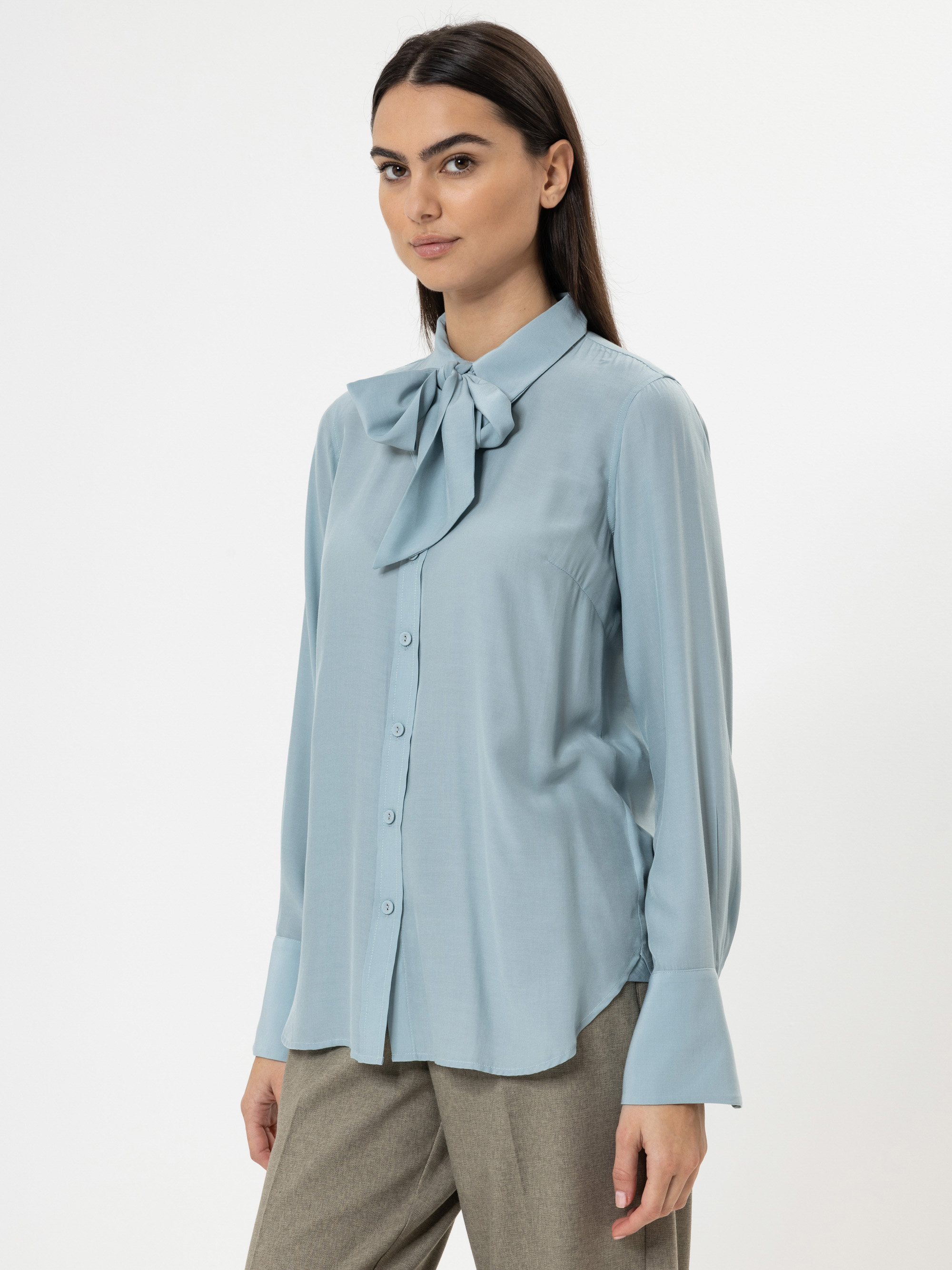 Eine Frau, die eine hellblaue Bluse mit einer großen Schleife am Kragen und breiten Manschetten sowie eine hellbraune Hose trägt, steht vor einem schlichten weißen Hintergrund.