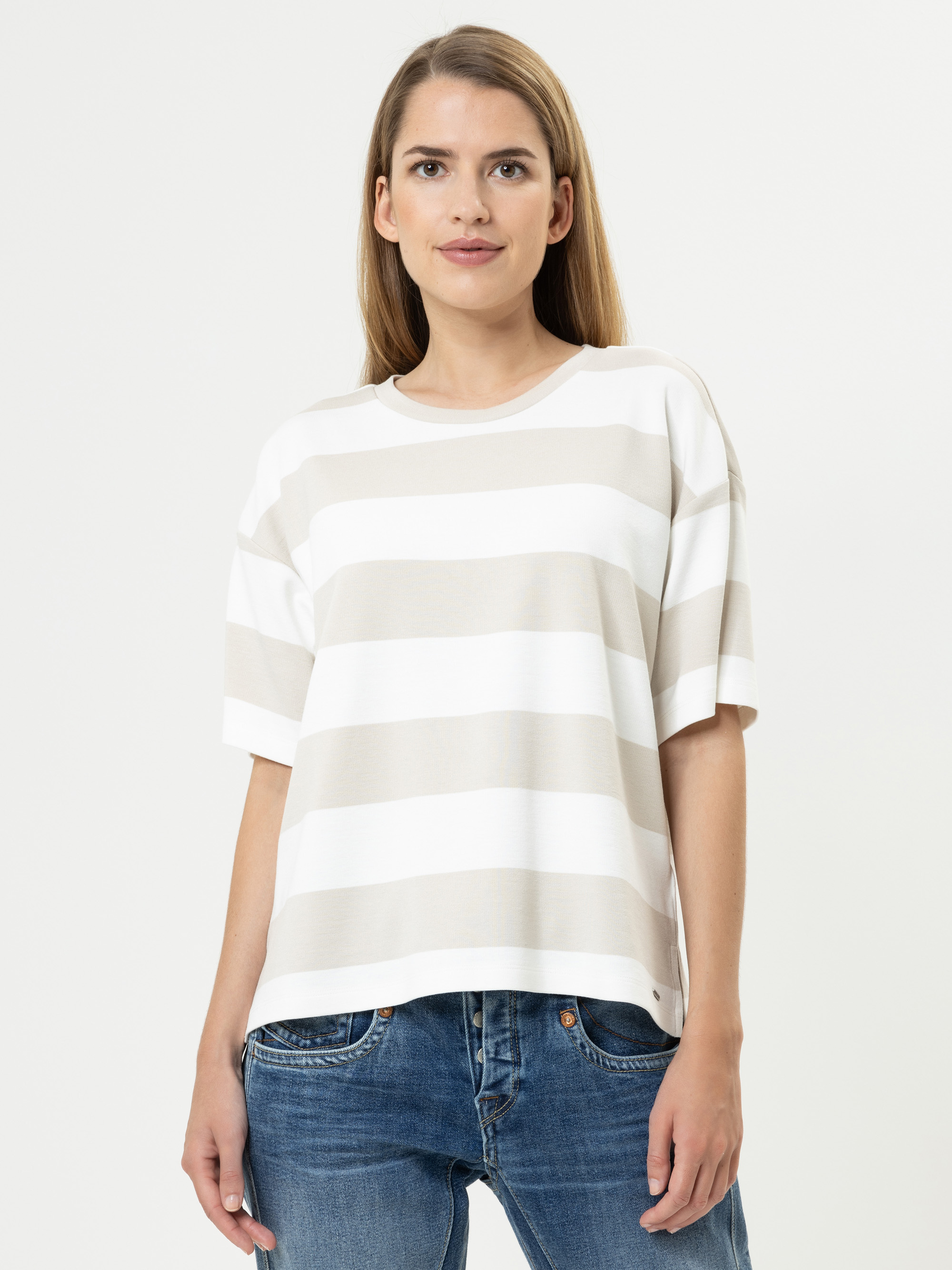 Eine Frau mit glattem, hellbraunem Haar trägt ein locker sitzendes, kurzärmeliges T-Shirt mit breiten beigen und weißen Querstreifen und blaue Jeans vor einem schlichten weißen Hintergrund.