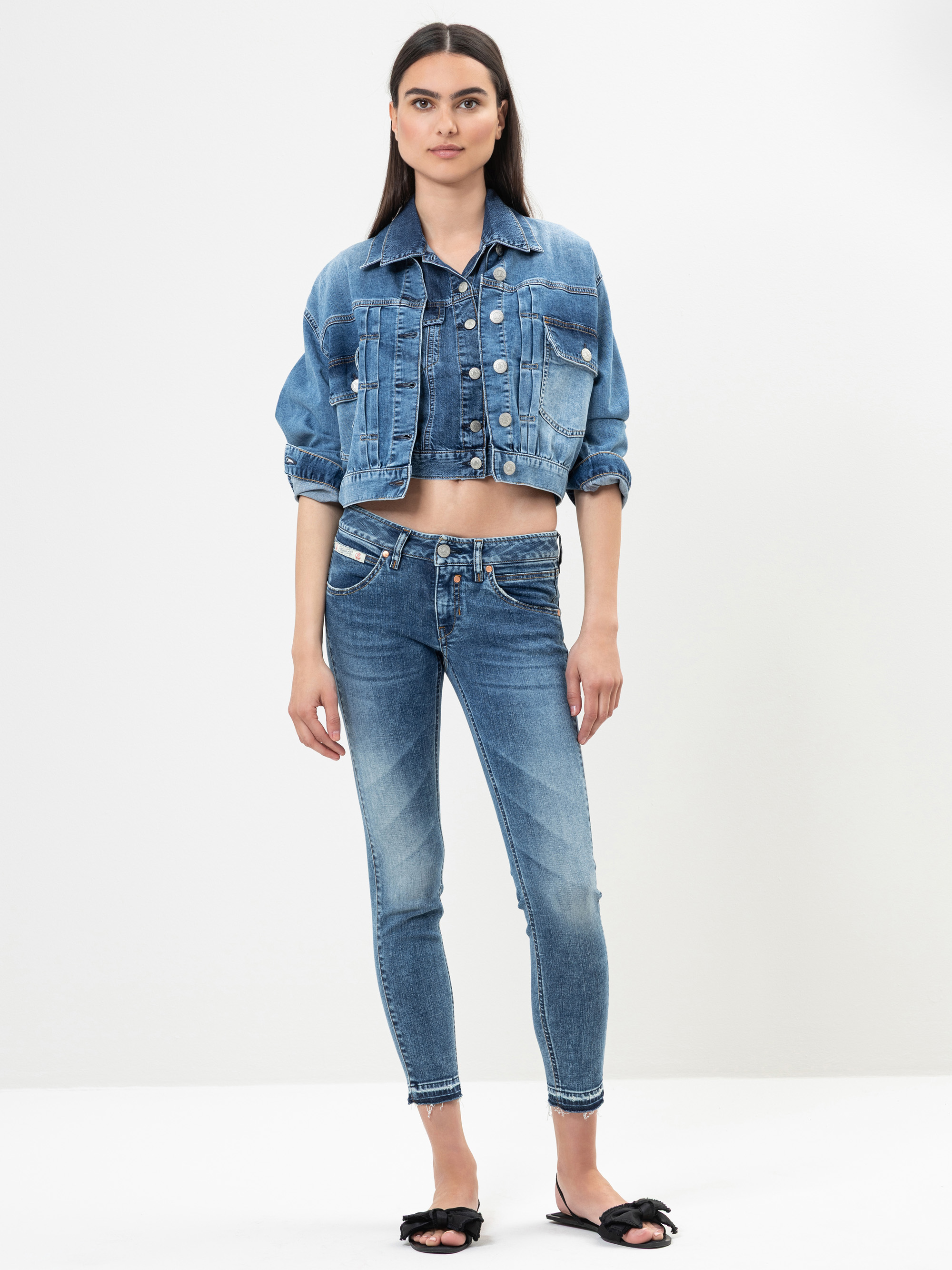 Eine Frau steht vor einem schlichten weißen Hintergrund und trägt eine blaue Jeansjacke, ein passendes Jeans-Crop-Top und Skinny Jeans. Sie ist barfuß, mit Ausnahme von schwarzen Slippersandalen mit Schleifen.