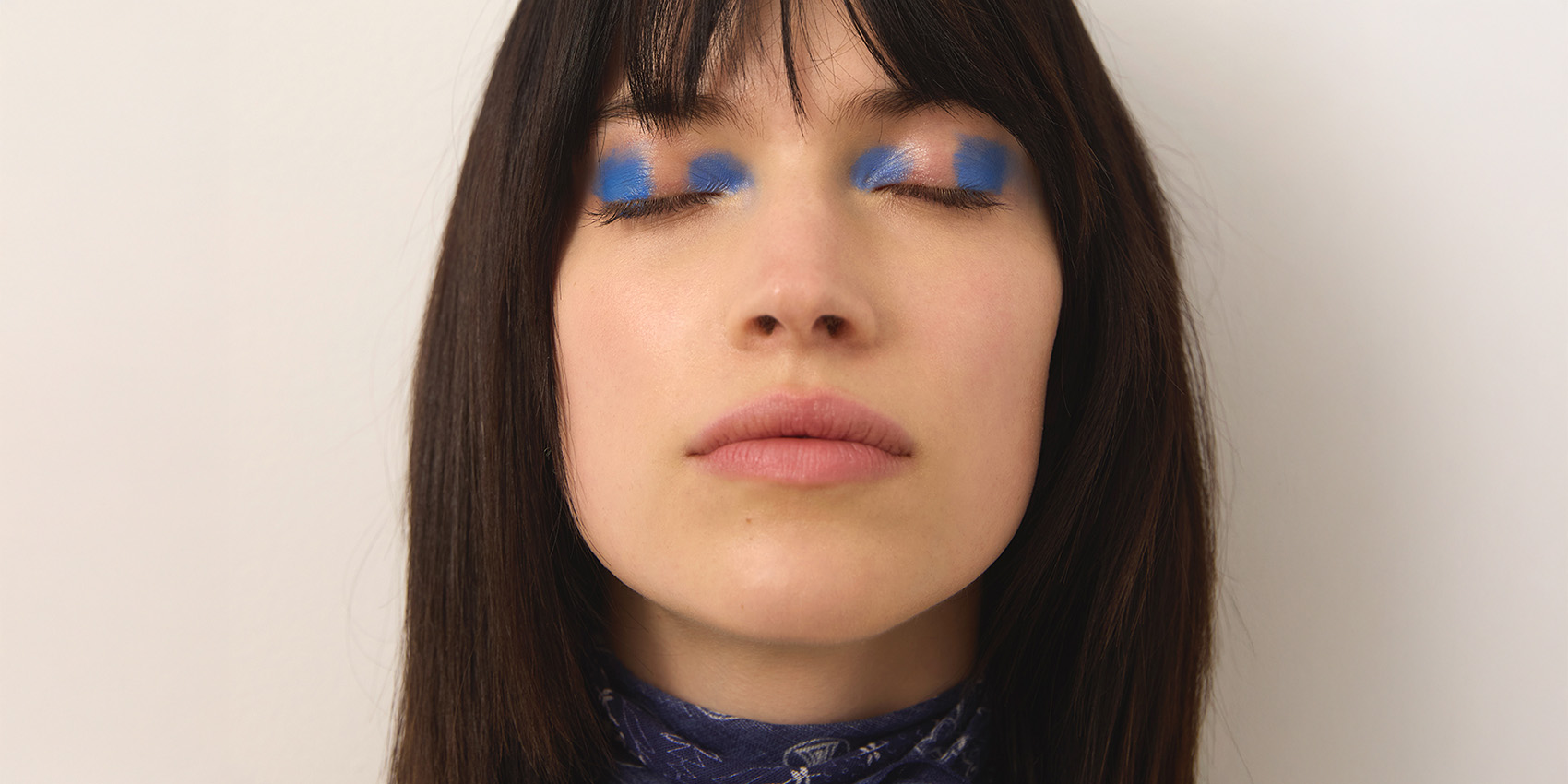 Eine Frau mit langem dunklem Haar und Pony blickt mit geschlossenen Augen nach vorne und trägt auf ihren Augenlidern kräftigen, hellblauen Lidschatten. Sie trägt einen gemusterten Schal um den Hals vor einem schlichten Hintergrund.