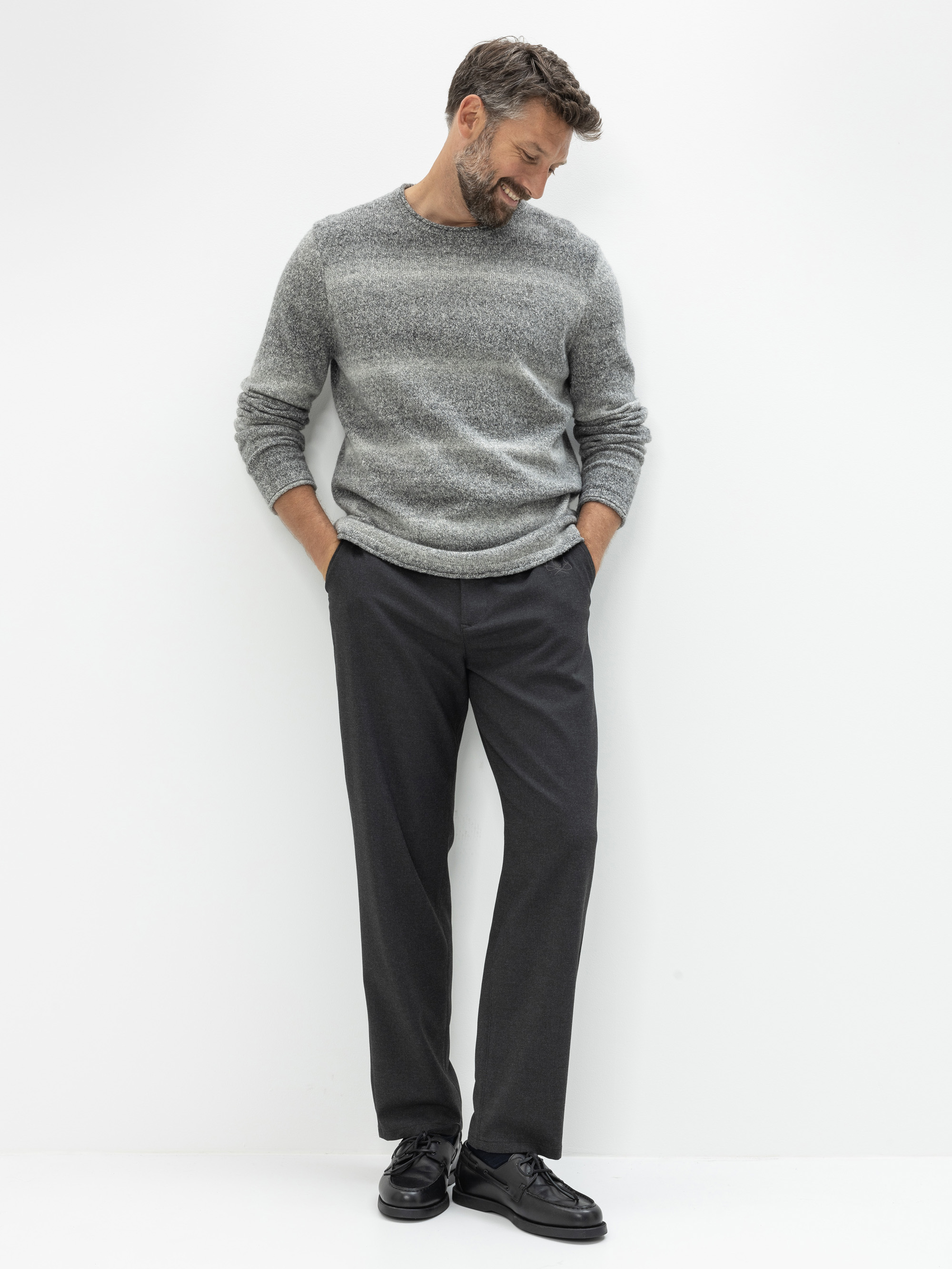 Ein Mann, der einen grauen Pullover, eine dunkle Hose und schwarze Schuhe trägt, steht vor einem schlichten weißen Hintergrund, blickt nach unten und lächelt, die Hände in den Taschen.