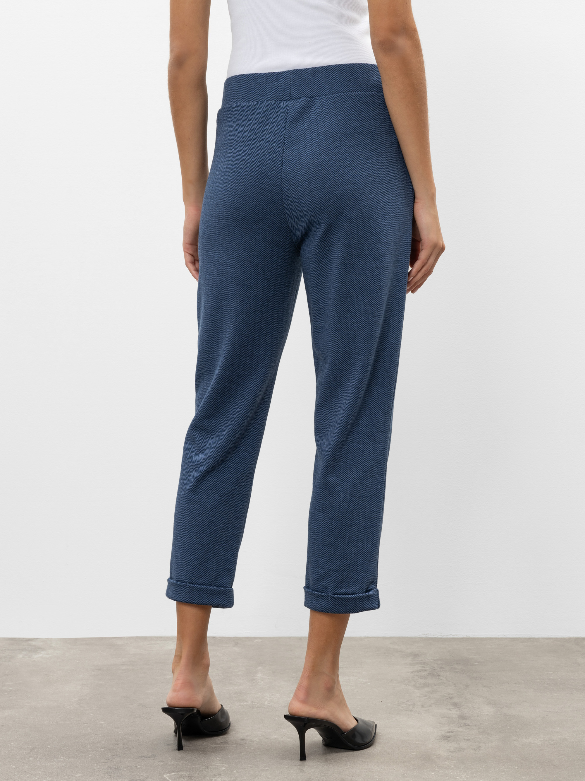 Eine Frau steht auf einem Betonboden, von der Kamera abgewandt, und trägt ein weißes Tanktop, eine blaue knöchellange Hose mit aufgerollten Manschetten und schwarze Pantoffeln mit Absatz. Der Hintergrund ist eine schlichte weiße Wand.