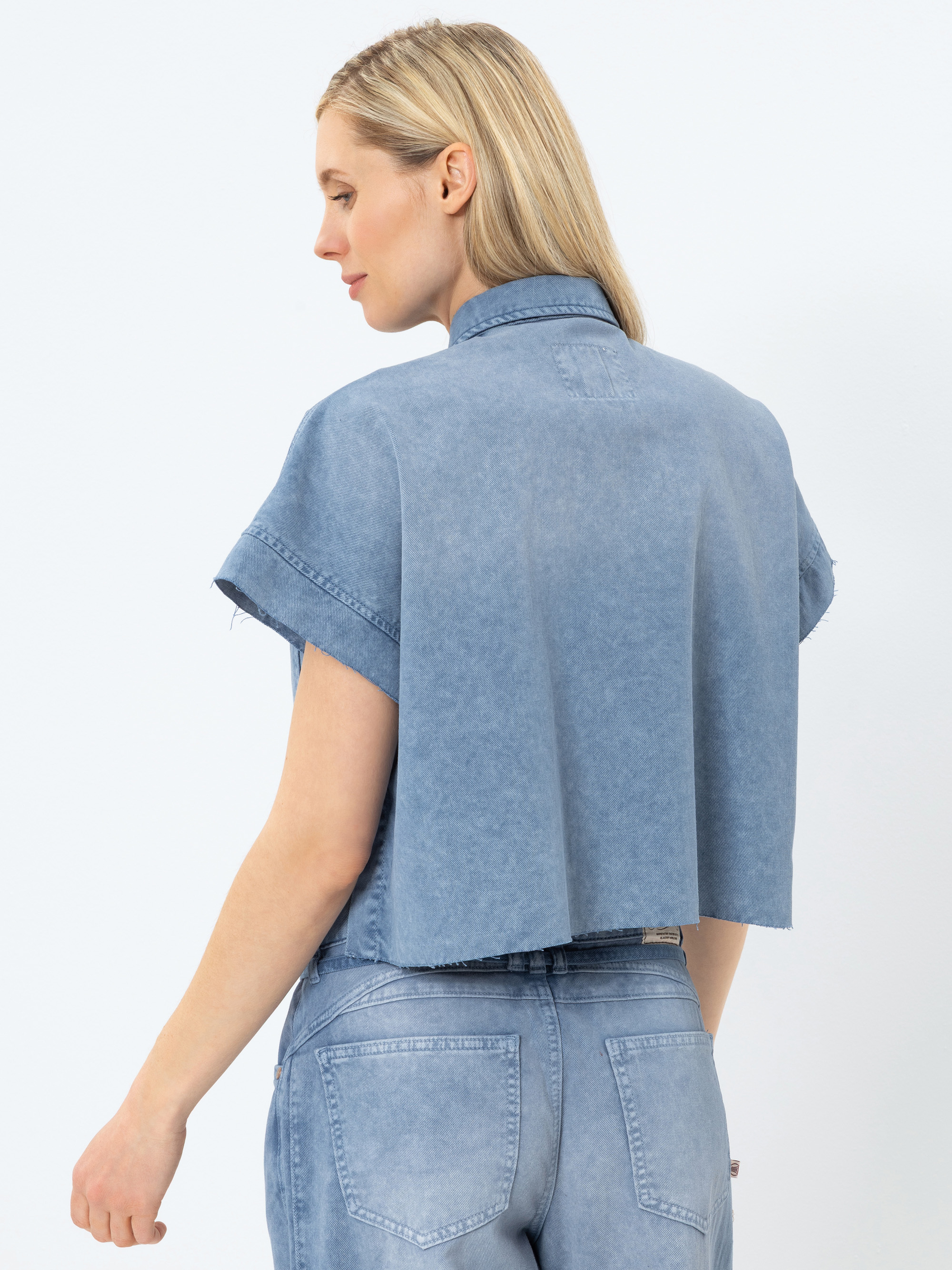 Eine Frau mit glattem blondem Haar ist von hinten zu sehen. Sie trägt ein kurzärmeliges, kurzgeschnittenes Jeanshemd und hellblaue Jeans mit hoher Taille. Sie steht vor einem schlichten weißen Hintergrund und blickt leicht zur Seite.