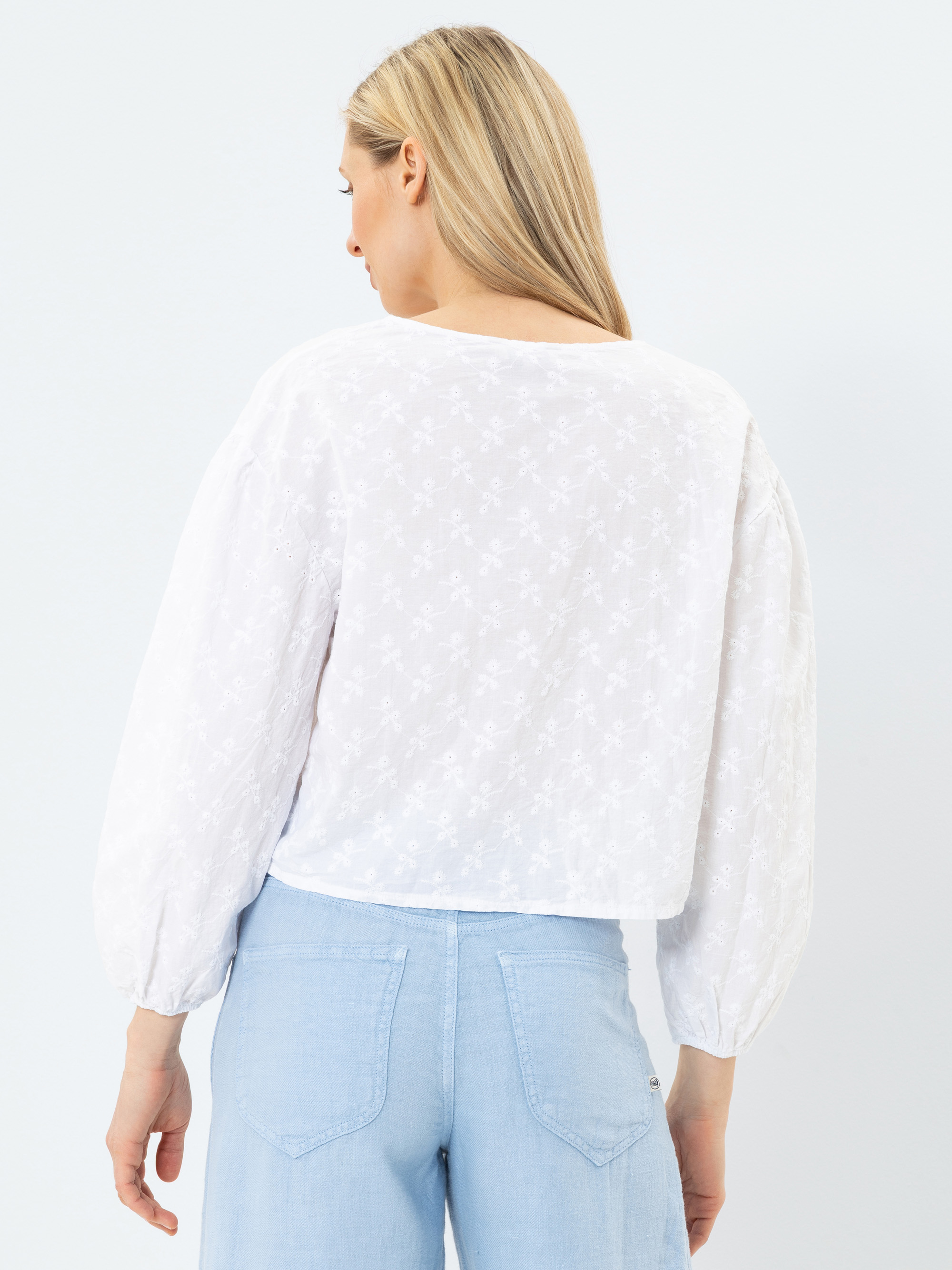 Eine Frau mit langen blonden Haaren, die eine weiße, langärmelige, bestickte Bluse und hellblaue, hochgekrempelte Jeans trägt, steht mit dem Rücken zur Kamera vor einem schlichten, hellen Hintergrund.