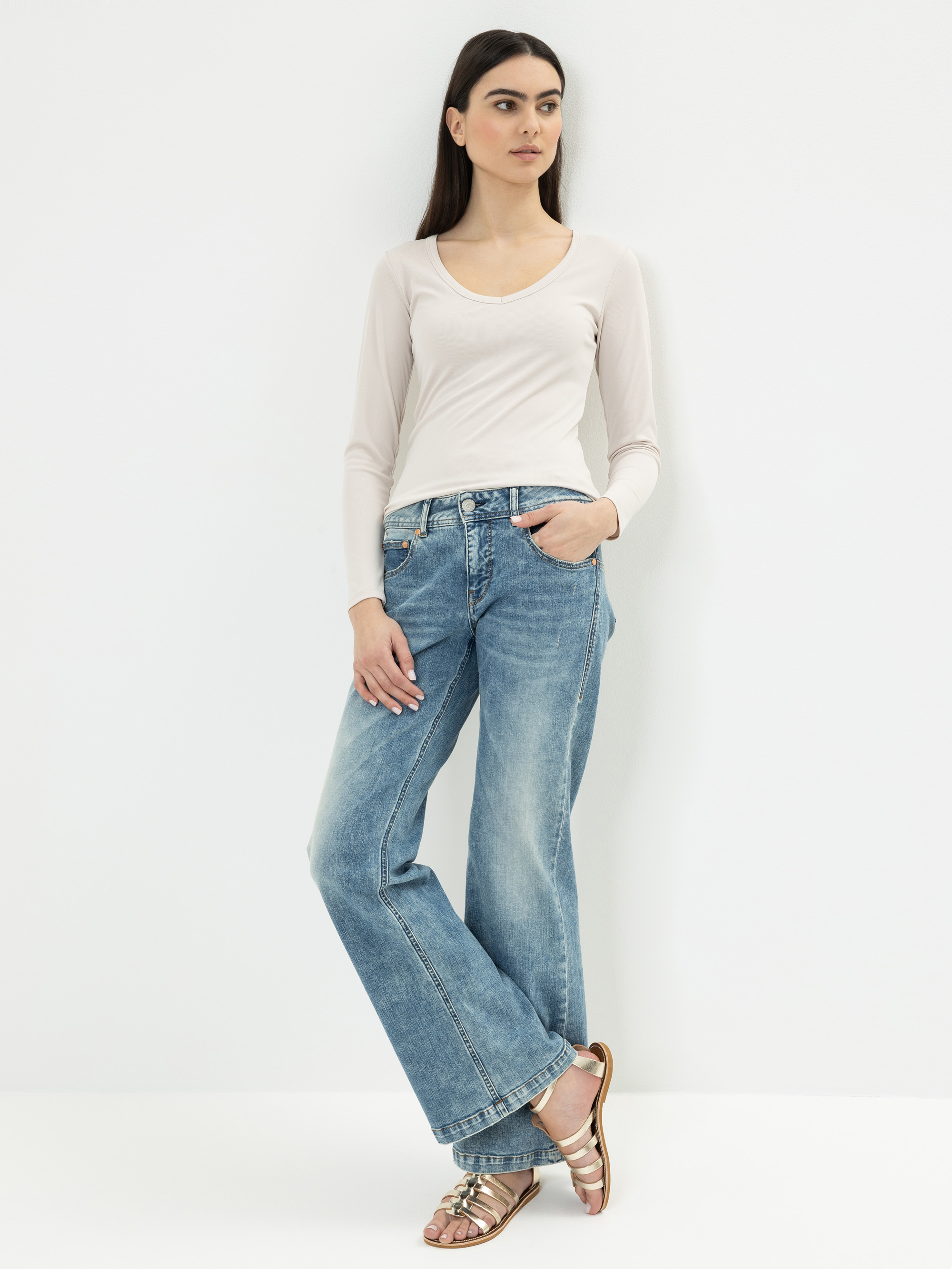 Eine Frau mit langen dunklen Haaren steht vor einer weißen Wand. Sie trägt ein hellbeiges, langärmeliges Oberteil, blaue Jeans mit weitem Bein und Metallic-Sandalen. Sie hat eine Hand in ihrer Tasche und schaut zur Seite.