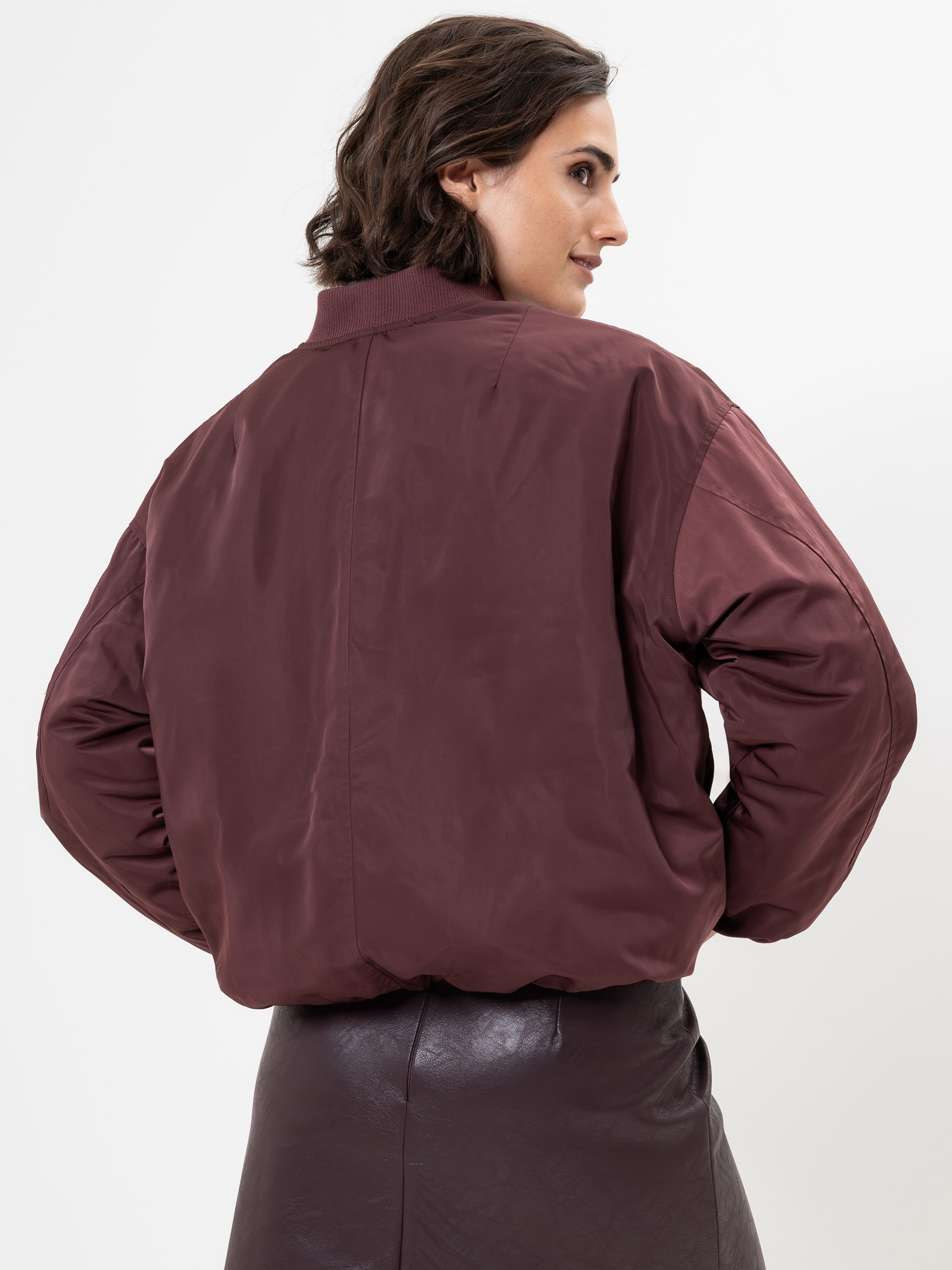 Eine Frau mit kurzen braunen Haaren blickt von sich weg. Sie trägt eine kastanienbraune Bomberjacke und einen dunklen Lederrock vor einem schlichten weißen Hintergrund.