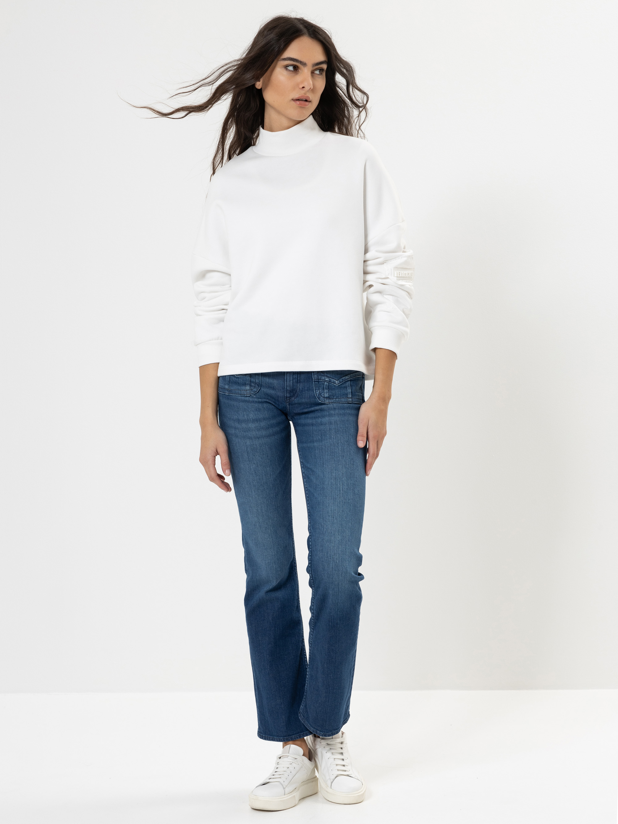 Eine Frau mit langen braunen Haaren, die einen weißen Rollkragenpullover, blaue Jeans und weiße Turnschuhe trägt, steht vor einem schlichten weißen Hintergrund.