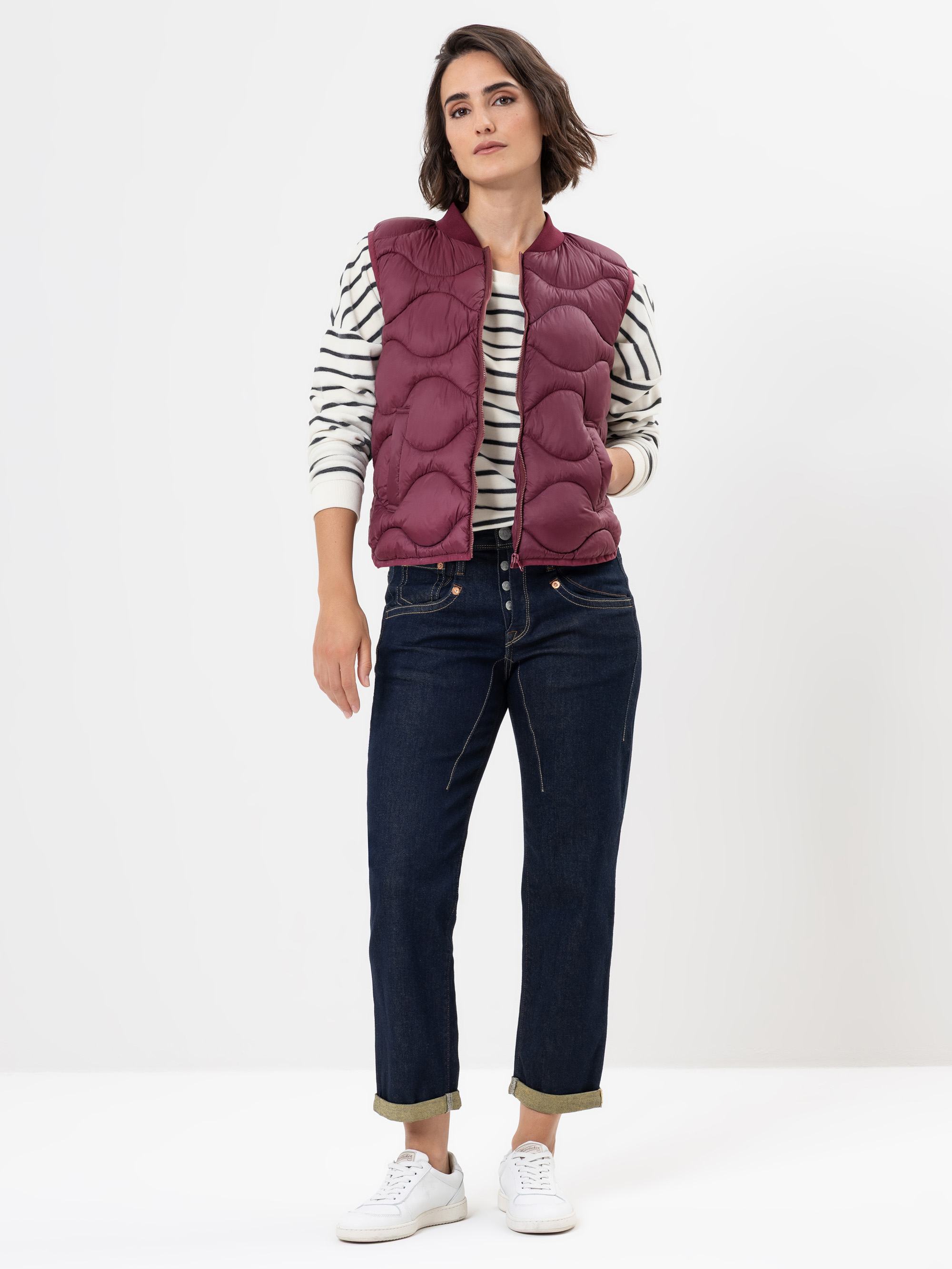 Eine Person steht vor einem weißen Hintergrund und trägt eine kastanienbraune Steppweste über einem weiß-schwarz gestreiften langärmeligen Hemd, dunkle Jeans mit Bündchen und weiße Turnschuhe.