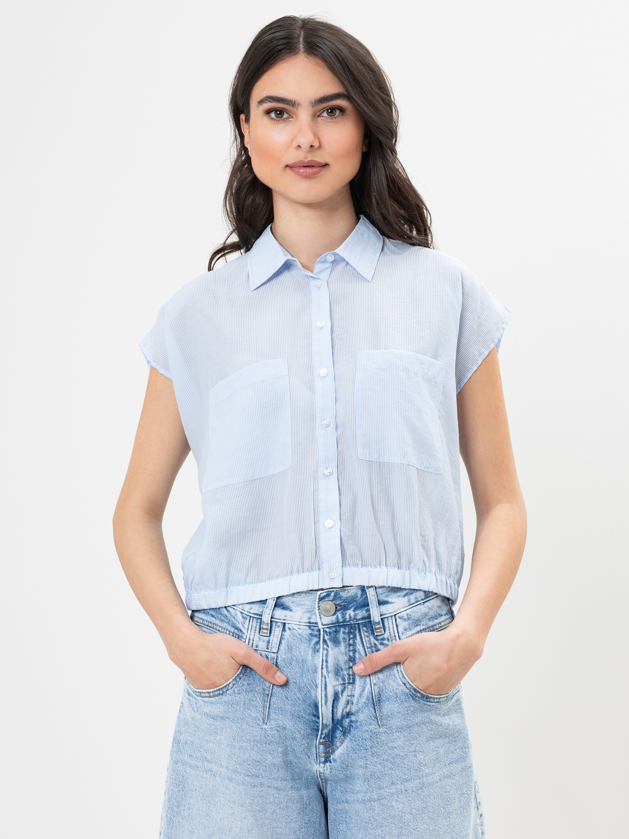 Eine Frau, die eine hellblaue, kurzärmelige, hochgeknöpfte Bluse mit zwei großen Vordertaschen und hellblaue Jeans mit hoher Taille trägt, steht mit den Händen in den Taschen vor einem schlichten weißen Hintergrund.