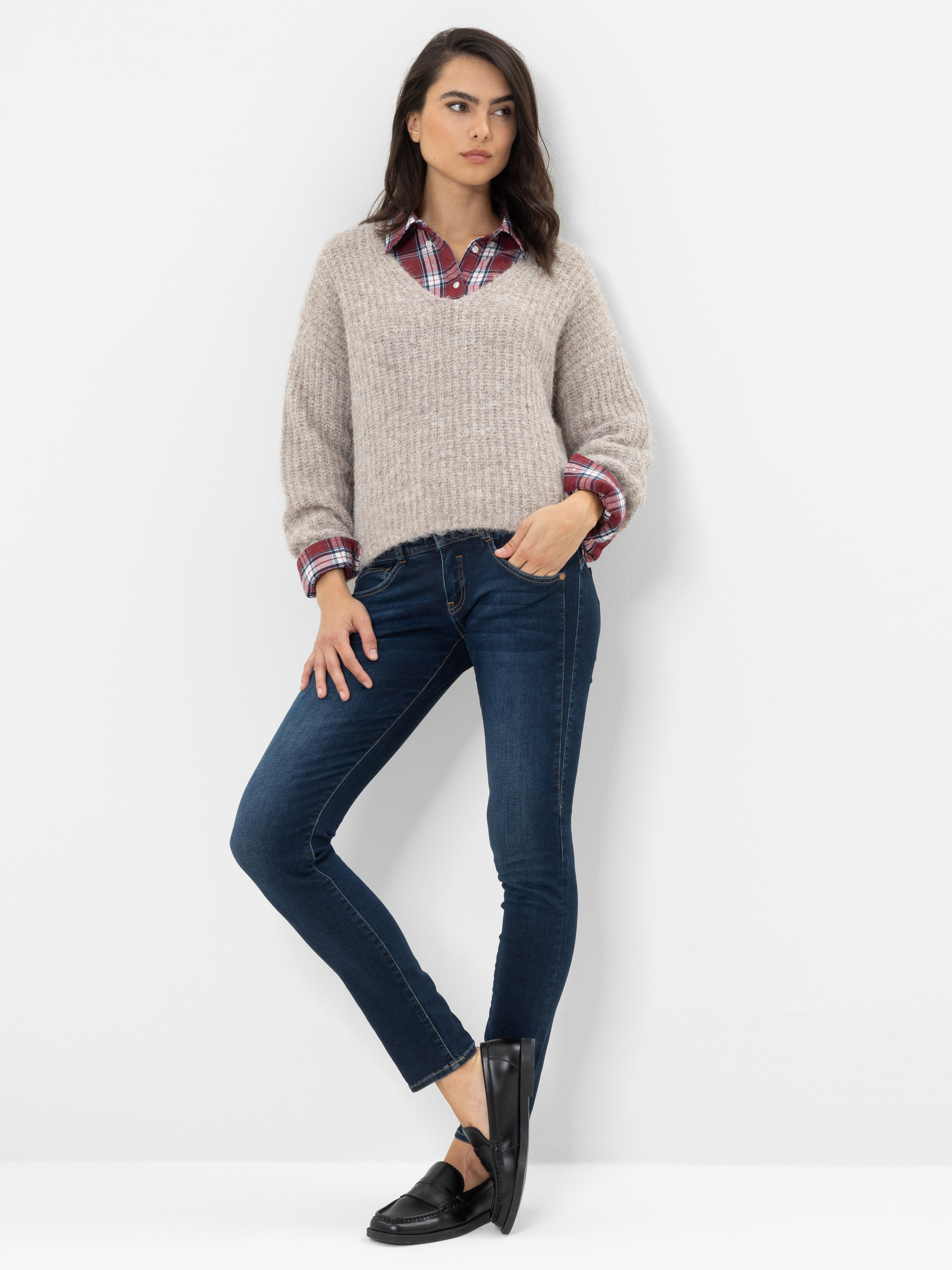 Eine Frau steht vor einer weißen Wand, trägt einen beigen Strickpullover über einem roten Karohemd, dunkelblaue Skinny-Jeans und schwarze Loafer, hat eine Hand in der Tasche und blickt zur Seite.