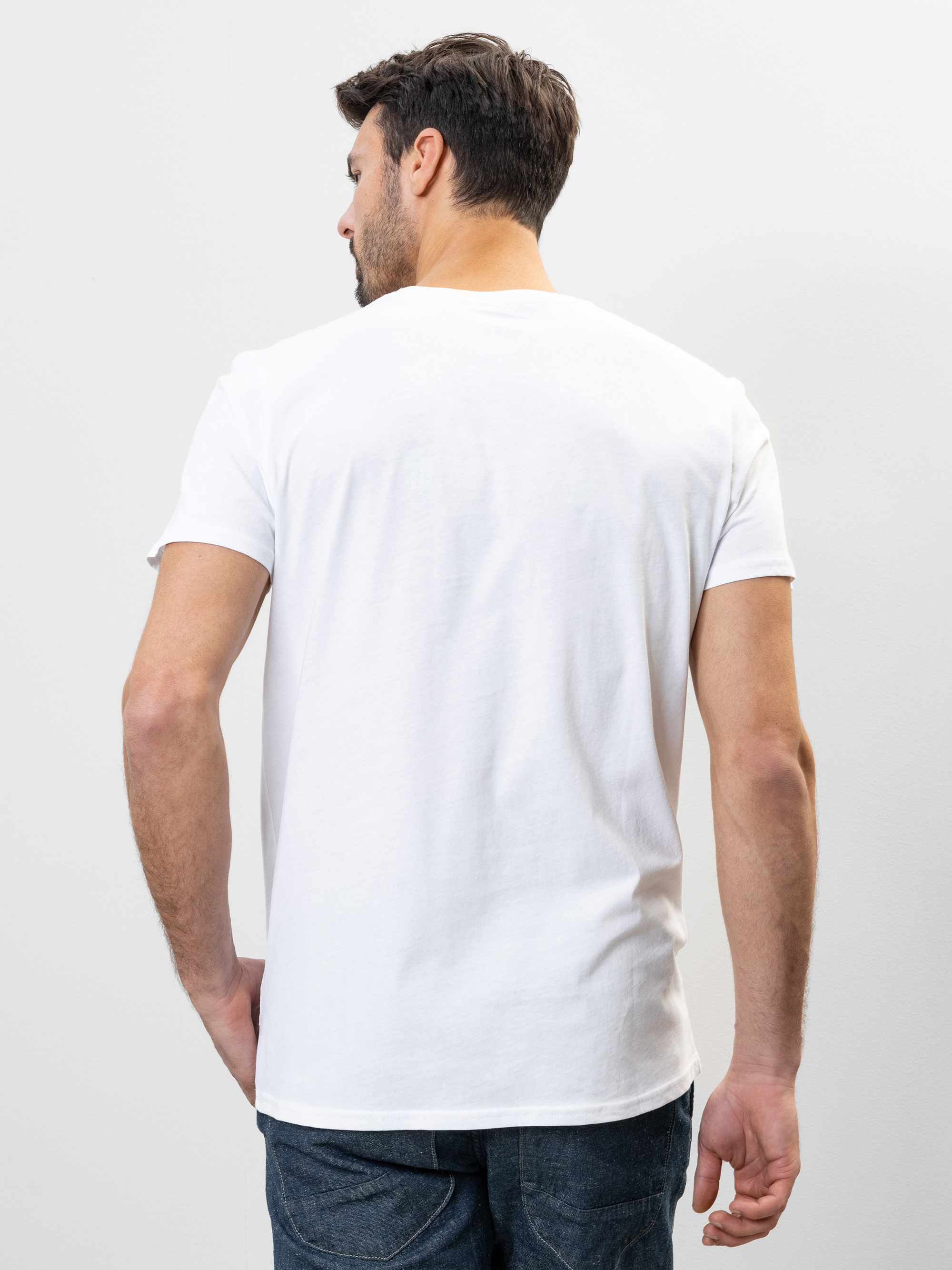 Ein Mann mit kurzen braunen Haaren steht mit dem Rücken zur Kamera, trägt ein schlichtes weißes T-Shirt und eine blaue Jeans vor einem schlichten weißen Hintergrund.