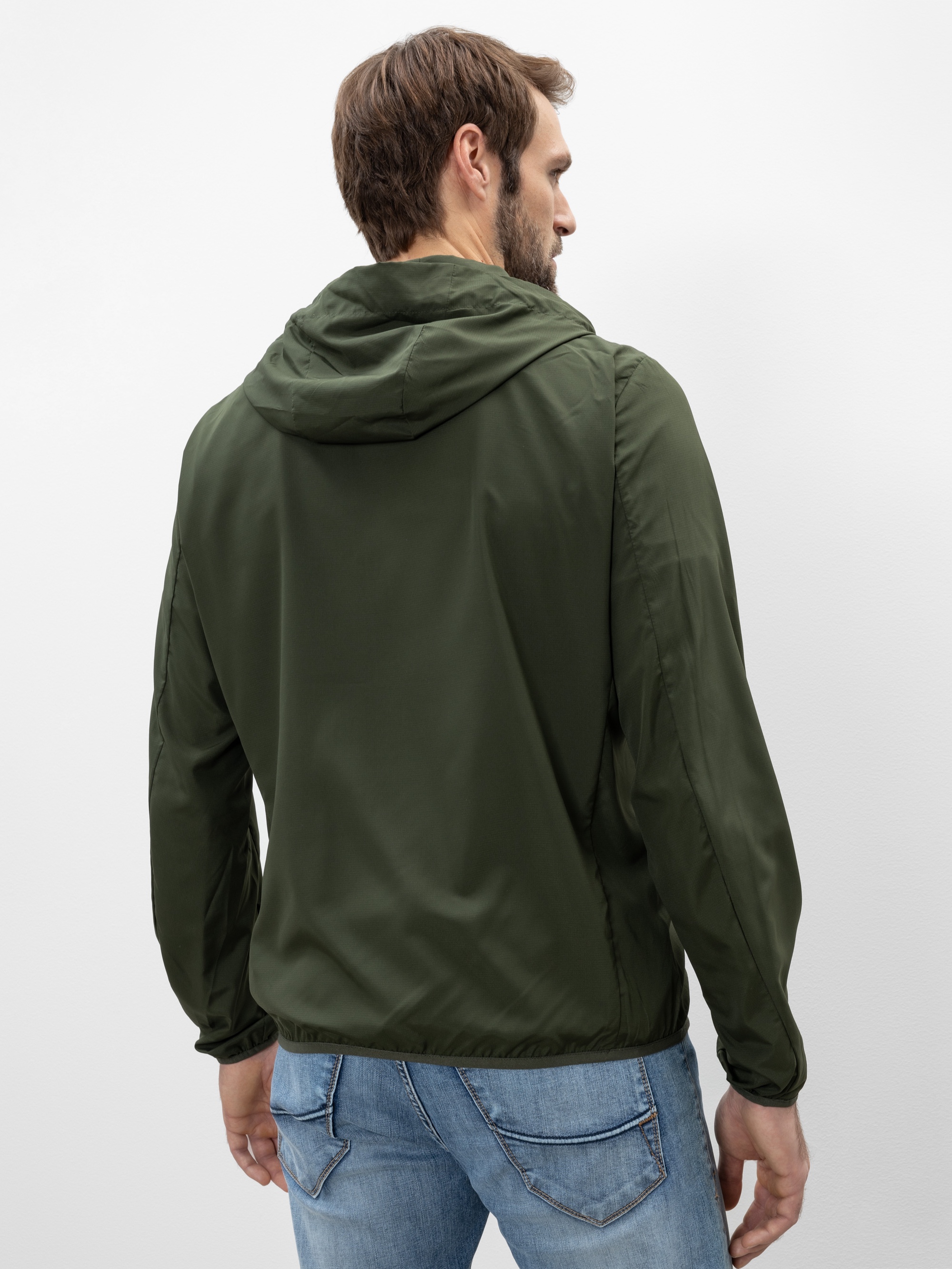 Ein Mann, der eine dunkelgrüne Kapuzenjacke und hellblaue Jeans trägt, steht mit dem Rücken zur Kamera vor einem schlichten weißen Hintergrund.
