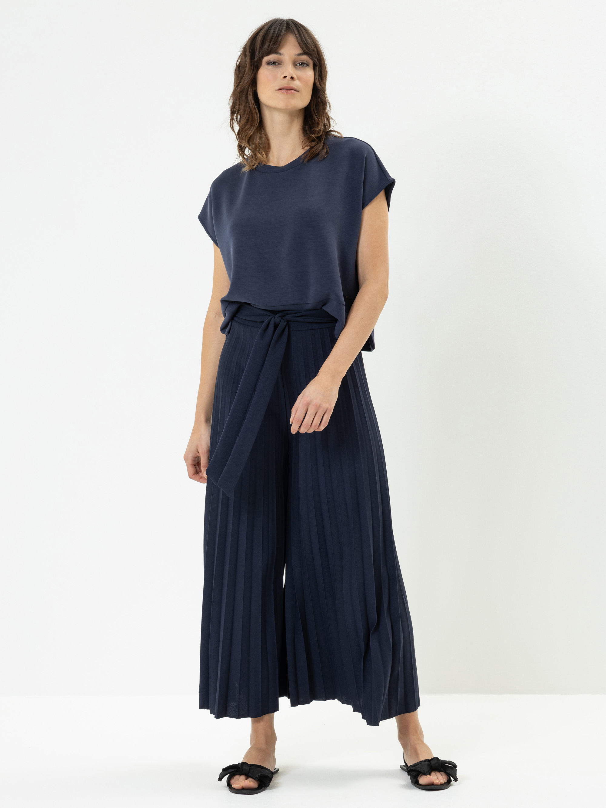 Eine Frau steht vor einem einfarbig weißen Hintergrund und trägt ein marineblaues, kurzärmeliges Oberteil und eine marineblaue Hose mit weitem Bein, die in Falten gelegt und mit einem passenden Stoffgürtel versehen ist, dazu schwarze, offene Schuhe.