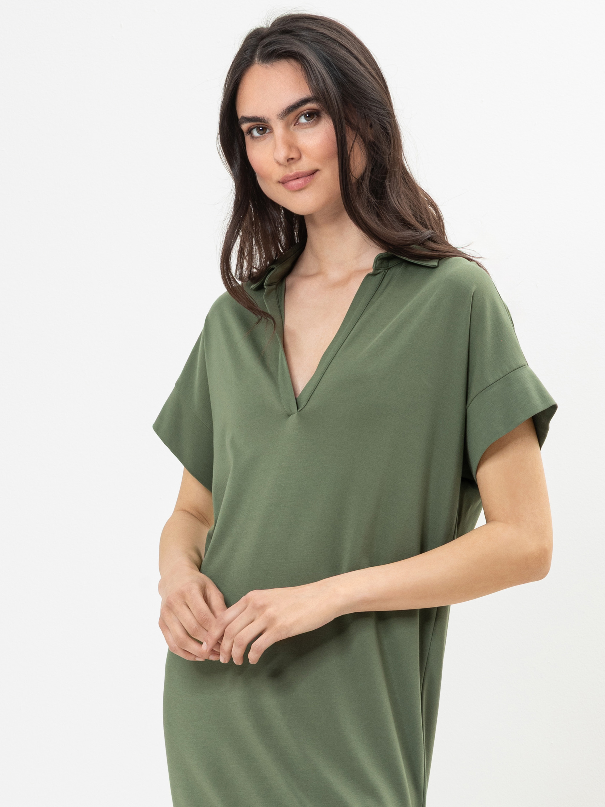 Eine Frau mit langen dunklen Haaren trägt ein olivgrünes, kurzärmeliges Kleid mit Kragen und V-Ausschnitt und steht vor einem schlichten weißen Hintergrund. Sie lächelt leicht und schaut in die Kamera.