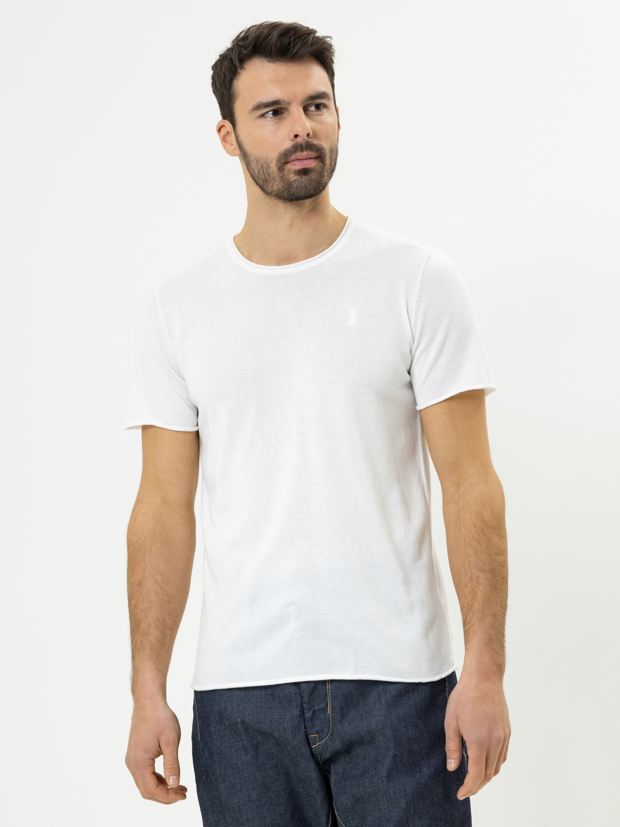 Ein Mann mit kurzen dunklen Haaren und einem Bart trägt ein einfaches weißes T-Shirt und dunkelblaue Jeans und steht vor einem einfachen weißen Hintergrund. Er blickt leicht zur Seite und hat einen neutralen Blick.