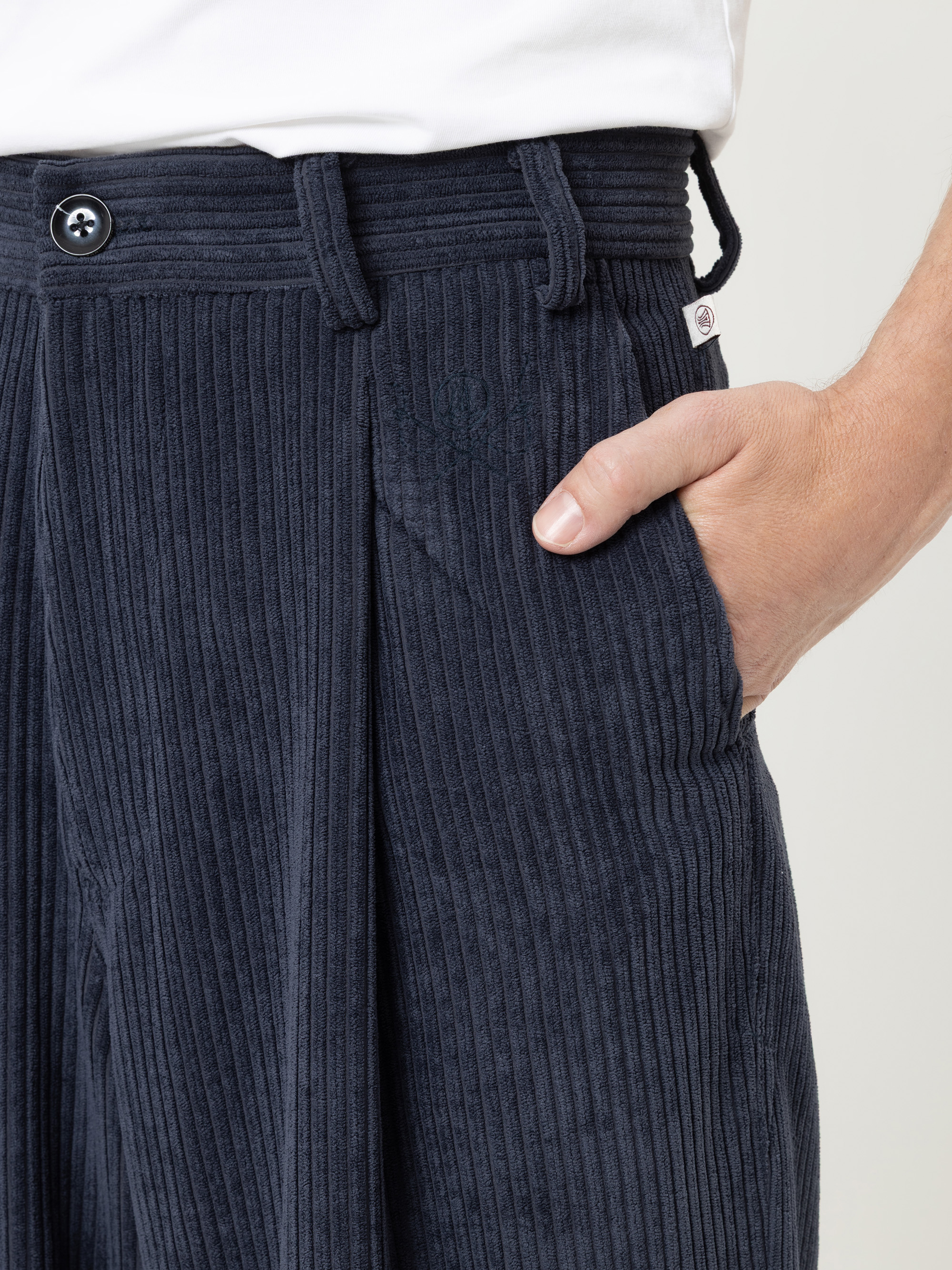 Eine Person, die eine dunkelblaue Cordhose und ein weißes Hemd trägt, hat die rechte Hand in der Tasche. Die Nahaufnahme zeigt die Struktur des Hosenstoffs, den Bund, die Gürtelschlaufen, den Knopf und ein kleines Logo über der Tasche.