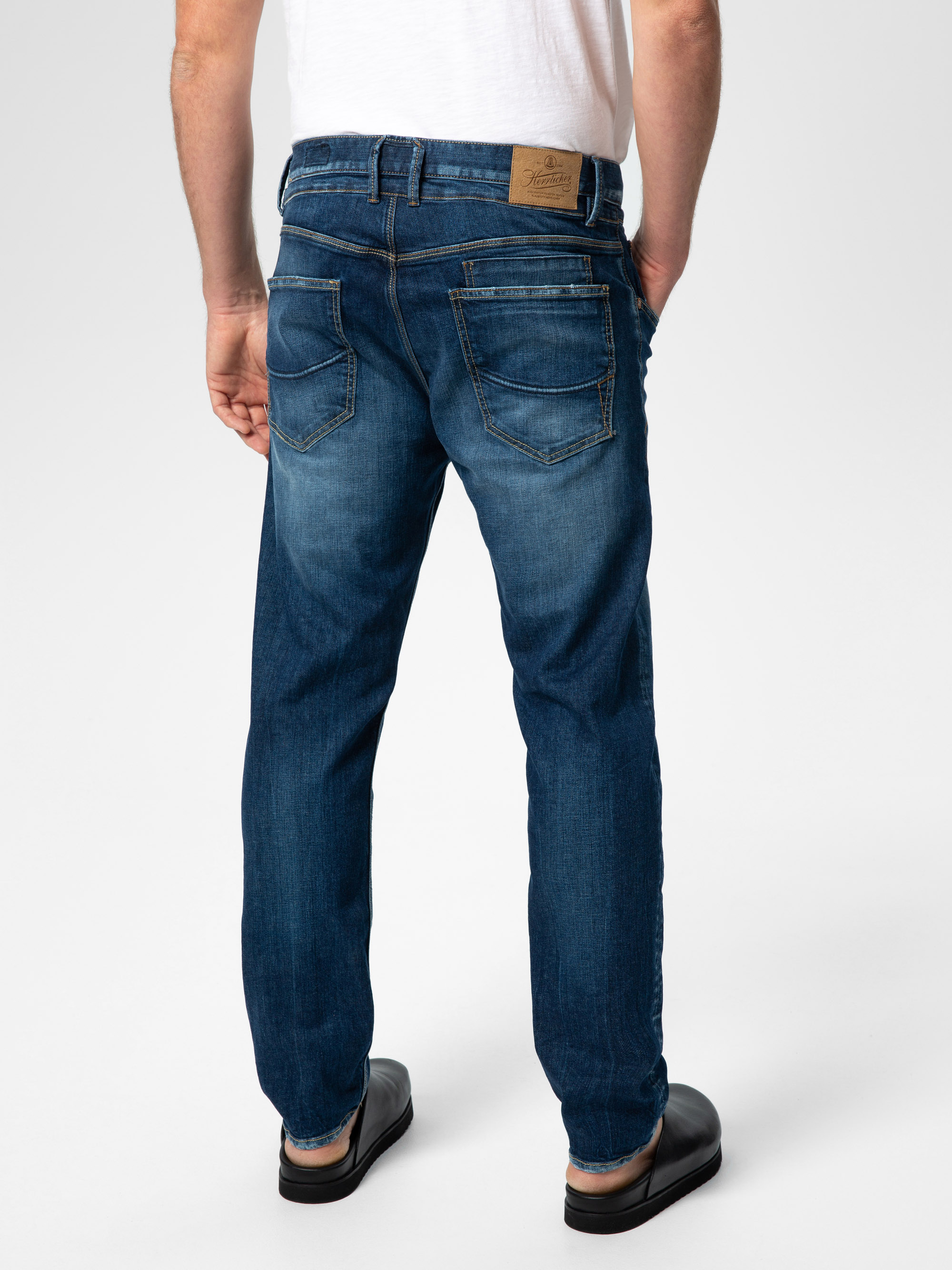 Eine Person, die eine dunkelblaue Jeans und schwarze Schuhe trägt, steht von der Kamera abgewandt und zeigt die Rückseite der Jeans mit sichtbaren Nähten und einem braunen Etikett am Bund. Die Person trägt außerdem ein weißes Hemd.