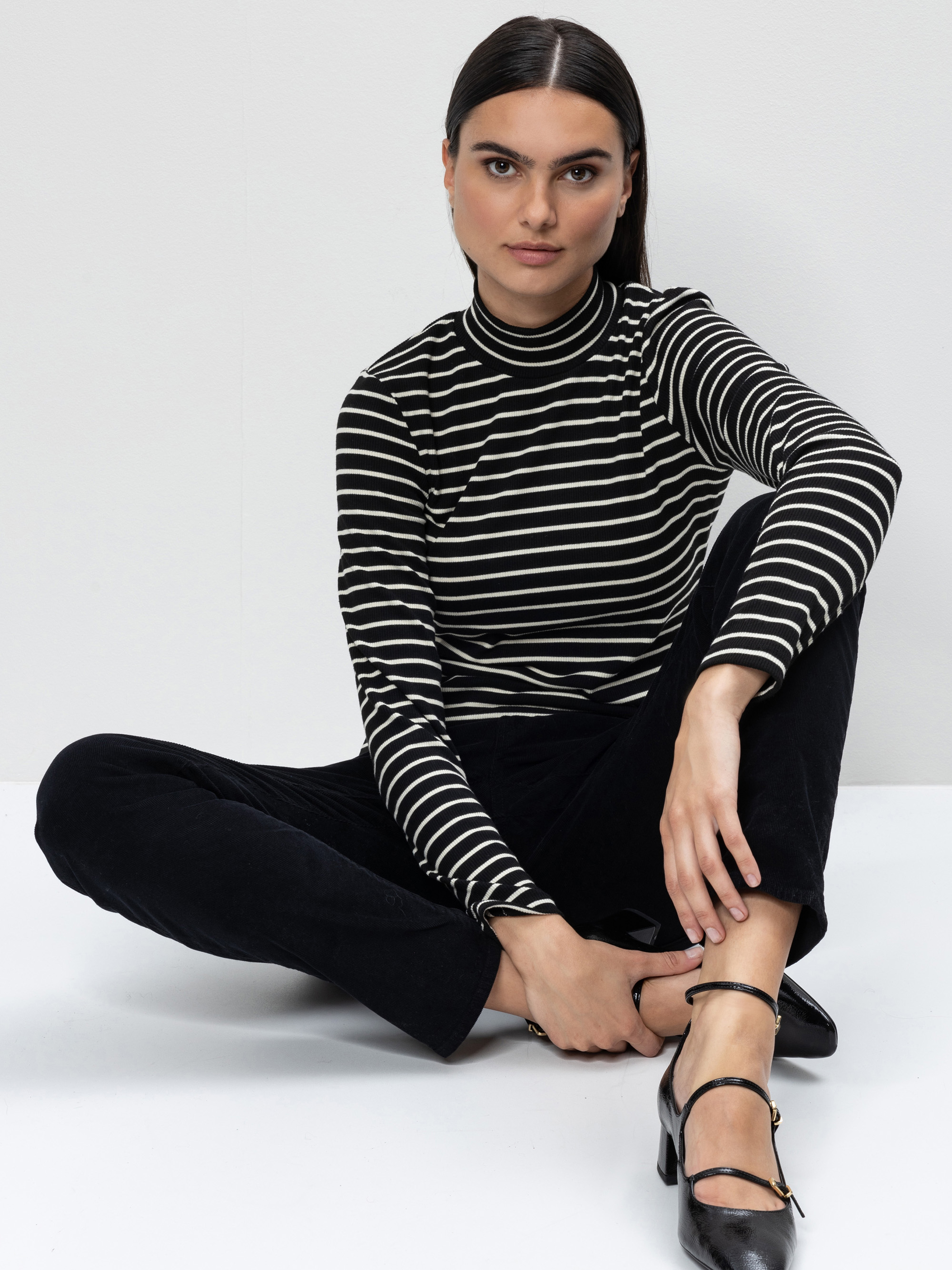 Eine Frau mit glattem dunklem Haar, die einen schwarz-weiß gestreiften Rollkragenpullover und eine schwarze Hose trägt, sitzt im Schneidersitz auf dem Boden vor einem schlichten weißen Hintergrund und schaut direkt in die Kamera. Sie trägt schwarze Ri