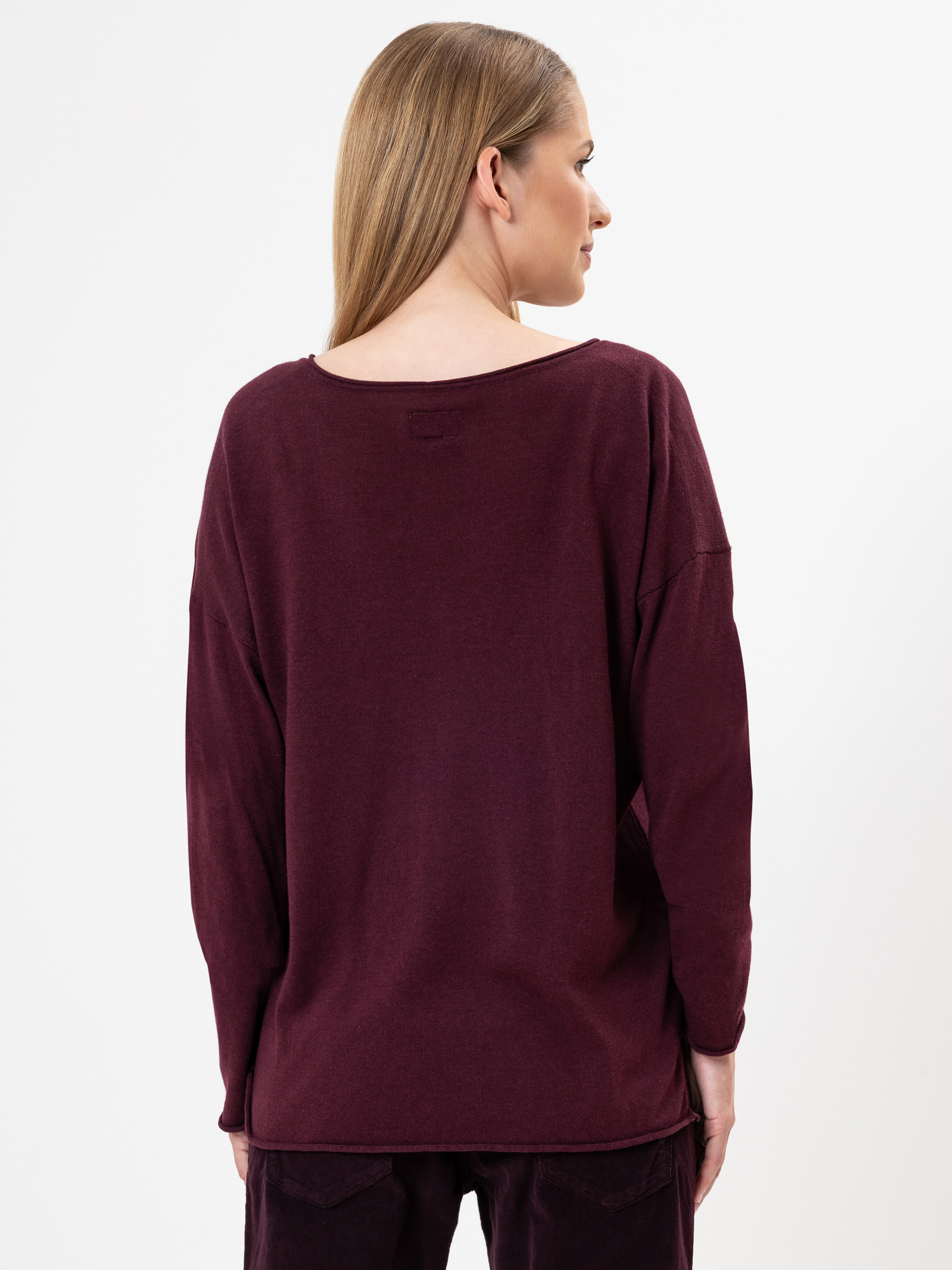 Eine Frau mit glattem, hellbraunem Haar steht mit dem Rücken zur Kamera und trägt ein locker sitzendes, langärmeliges kastanienbraunes Oberteil und eine dazu passende Hose vor einem schlichten weißen Hintergrund.