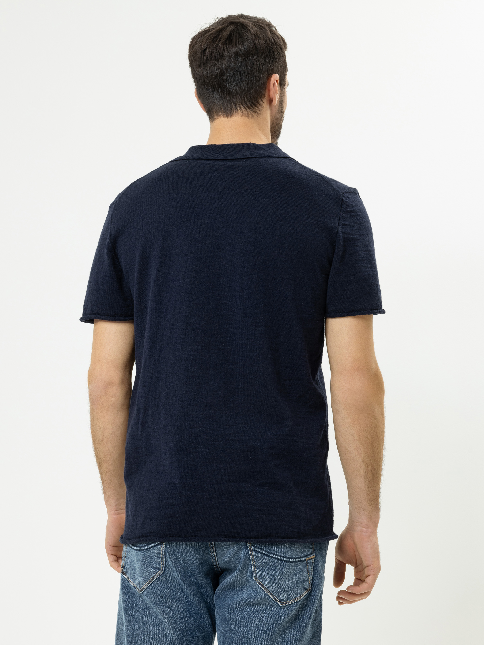 Ein Mann steht mit dem Rücken zur Kamera, trägt ein dunkles, marineblaues, kurzärmeliges T-Shirt und blaue Jeans vor einem schlichten weißen Hintergrund.