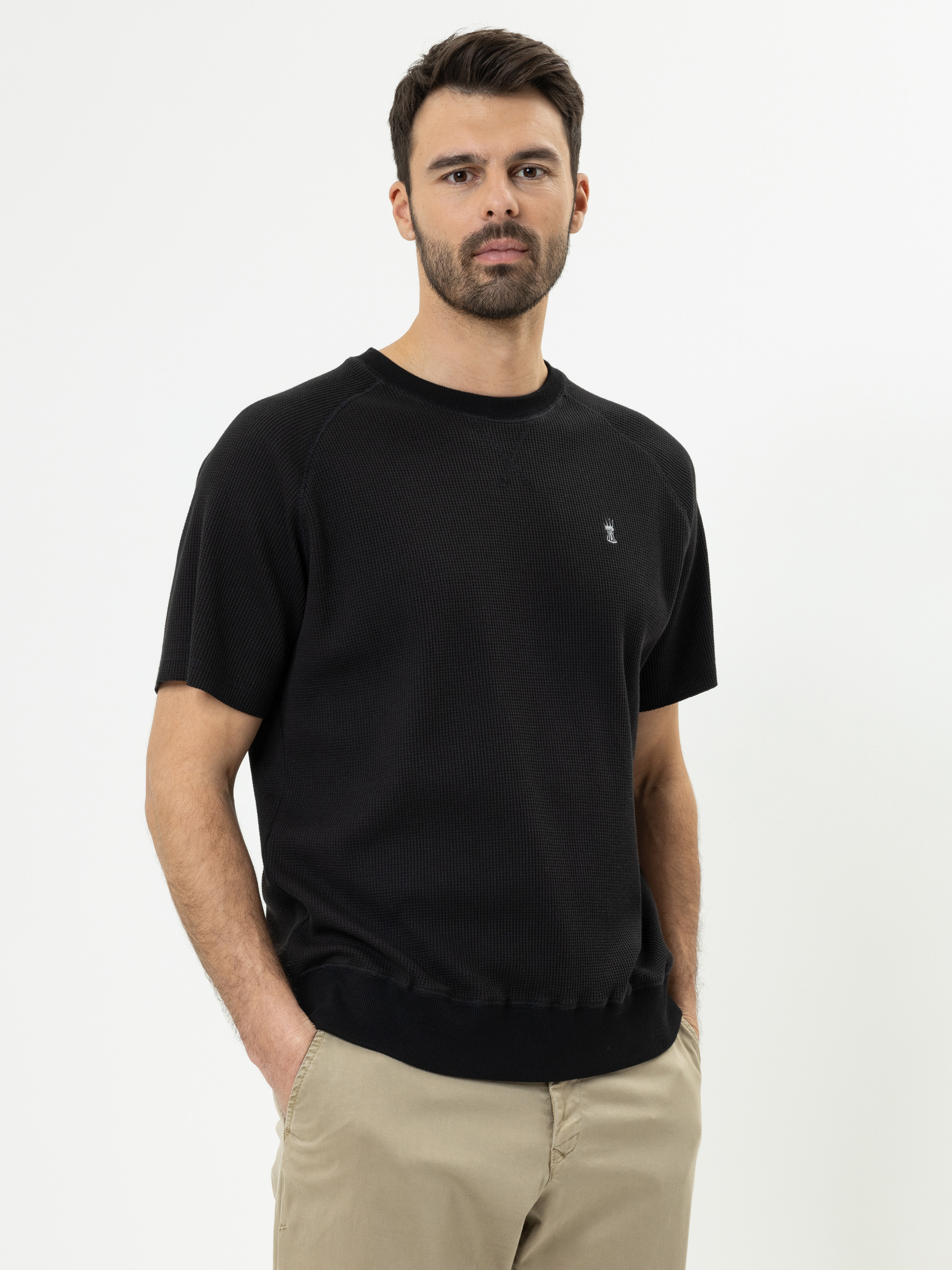 Ein Mann mit kurzen dunklen Haaren und einem Bart trägt ein schwarzes, kurzärmeliges T-Shirt mit Rundhalsausschnitt und eine beigefarbene Hose und steht vor einem schlichten weißen Hintergrund.