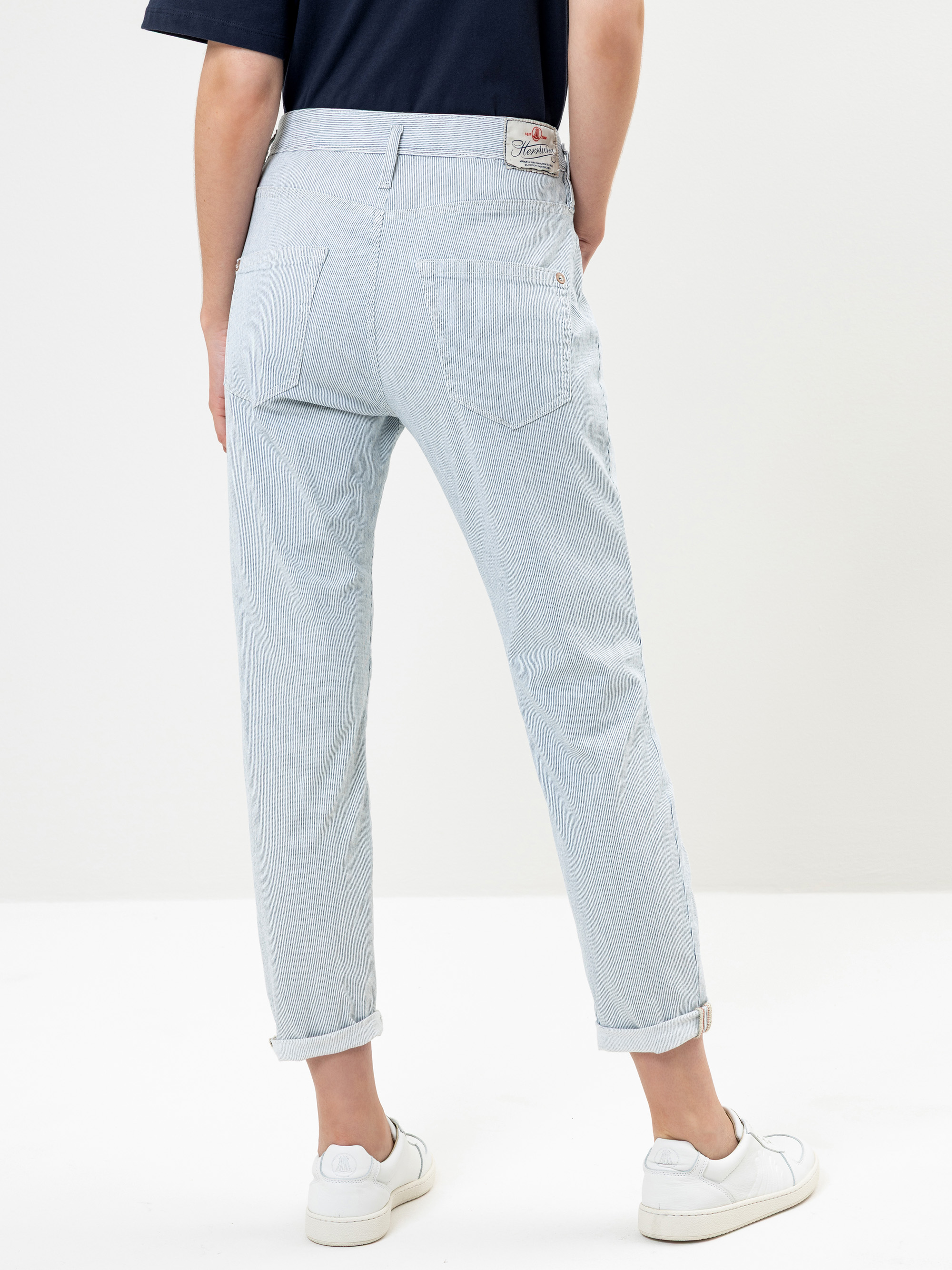 Mit dem Rücken zur Kamera stehende Person, die eine hellblau gestreifte Jeans mit aufgerollten Manschetten, ein marineblaues Hemd und weiße Turnschuhe vor einem schlichten weißen Hintergrund trägt.