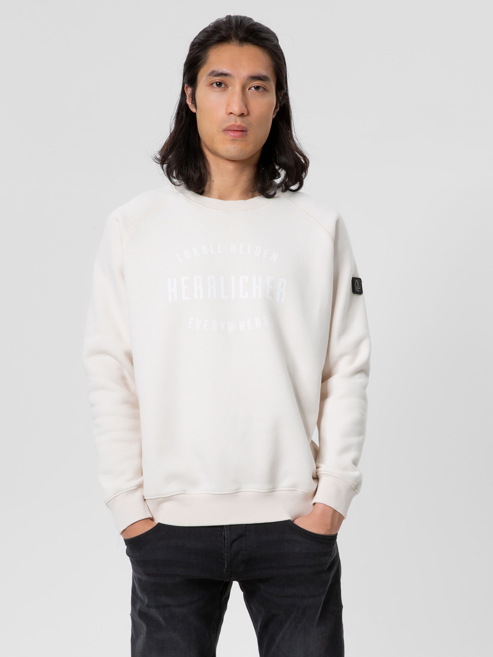 Eine Person mit langen dunklen Haaren trägt ein cremefarbenes Sweatshirt mit dem weißen Aufdruck Lokale Helden Herzlichkeit Everywhere, das vor einem schlichten hellgrauen Hintergrund steht, die Hände in den Taschen.