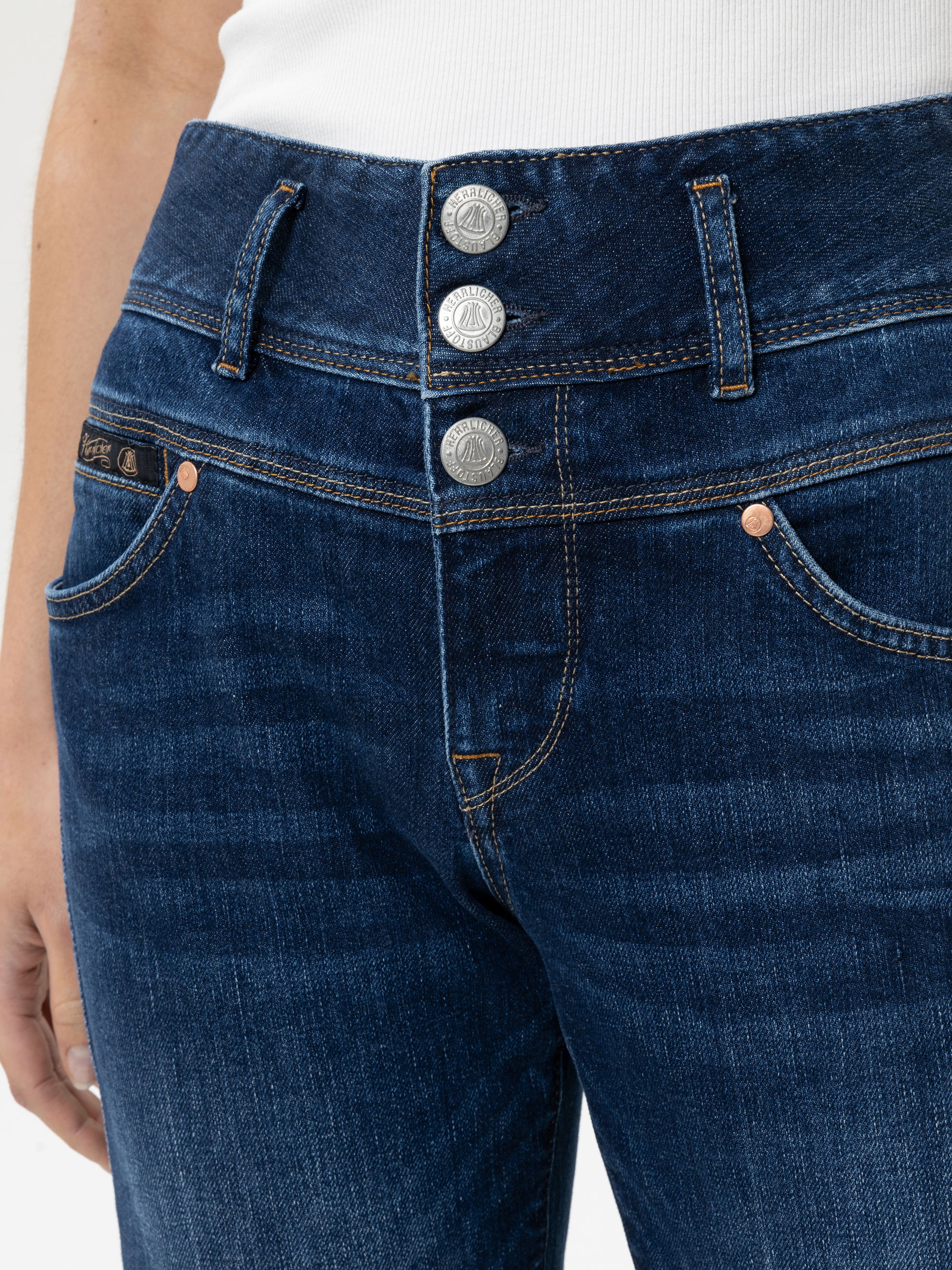 Nahaufnahme einer Person, die eine dunkelblaue Jeans mit drei silbernen Knöpfen und orangefarbenen Nähten trägt, dazu ein weißes geripptes Oberteil. Man sieht die Hand der Person, die an der Seite ruht.