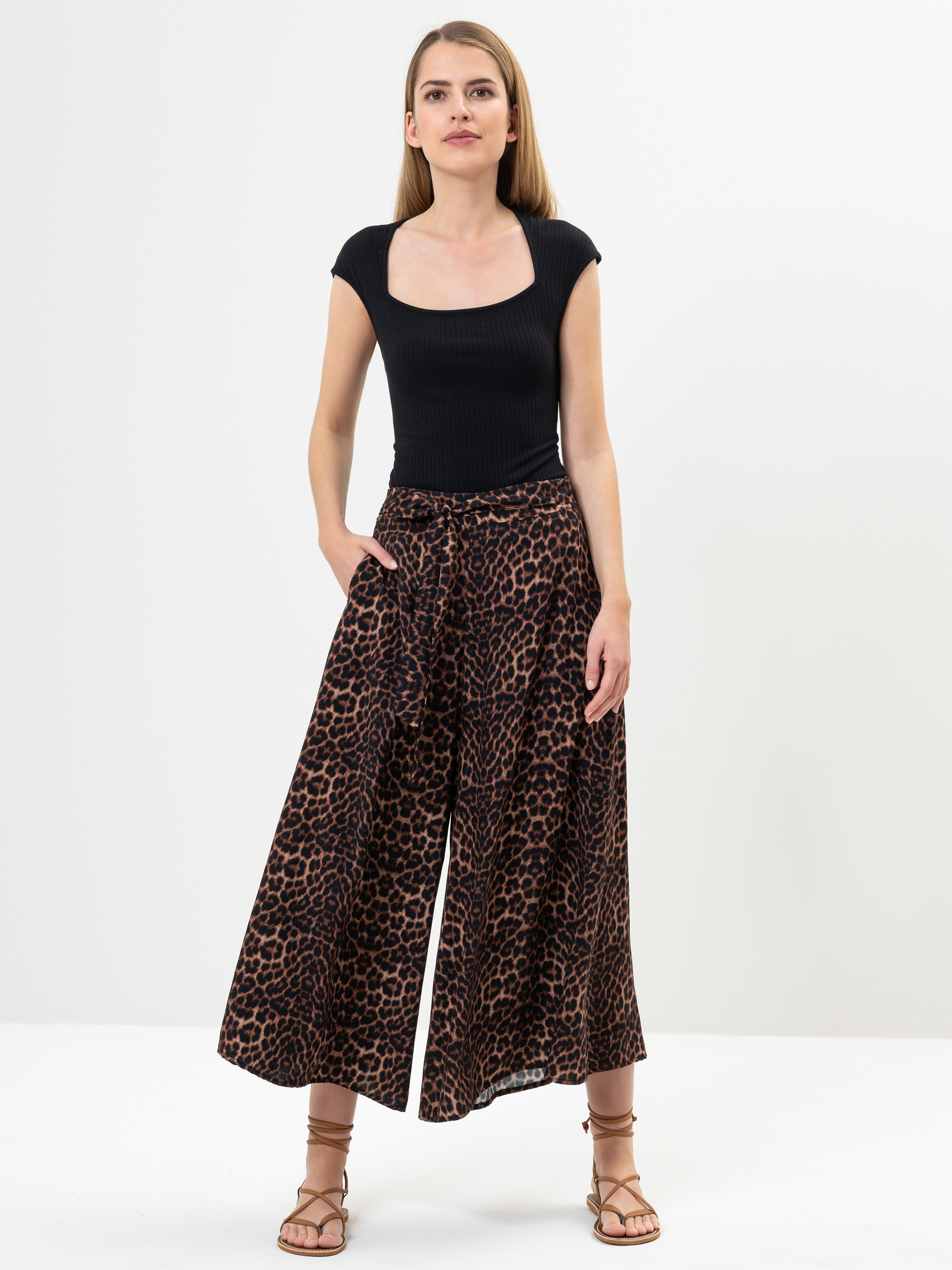 Eine Frau steht vor einem schlichten weißen Hintergrund, trägt ein schwarzes, kurzärmeliges Oberteil mit quadratischem Halsausschnitt, eine Hose mit weitem Bein und Leopardenmuster sowie hellbraune Riemchensandalen, eine Hand in der Tasche und einen ne