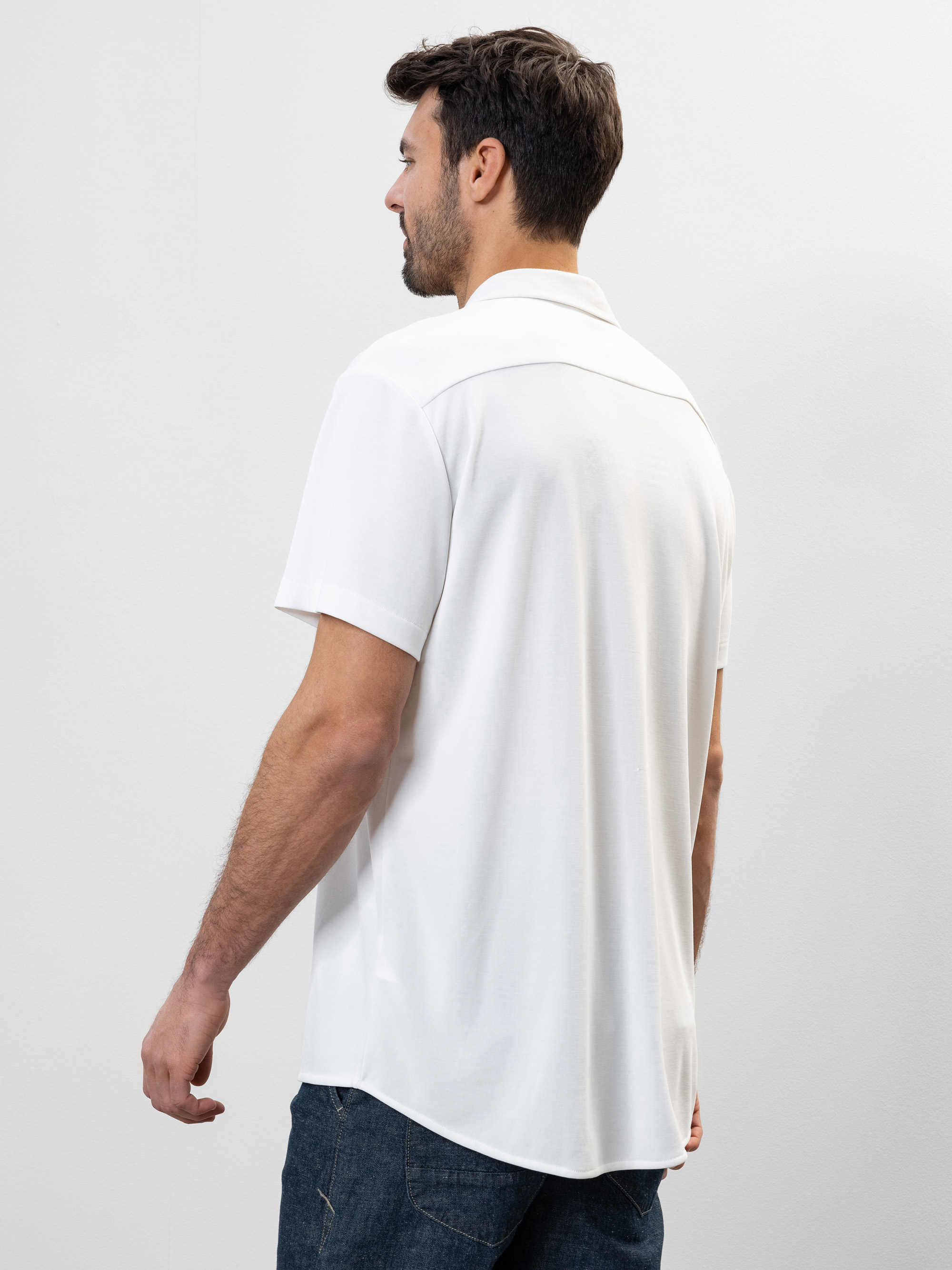 Ein Mann mit kurzen dunklen Haaren, der ein weißes kurzärmeliges Hemd und eine dunkle Jeans trägt, steht mit dem Gesicht zur Kamera vor einem schlichten hellen Hintergrund.