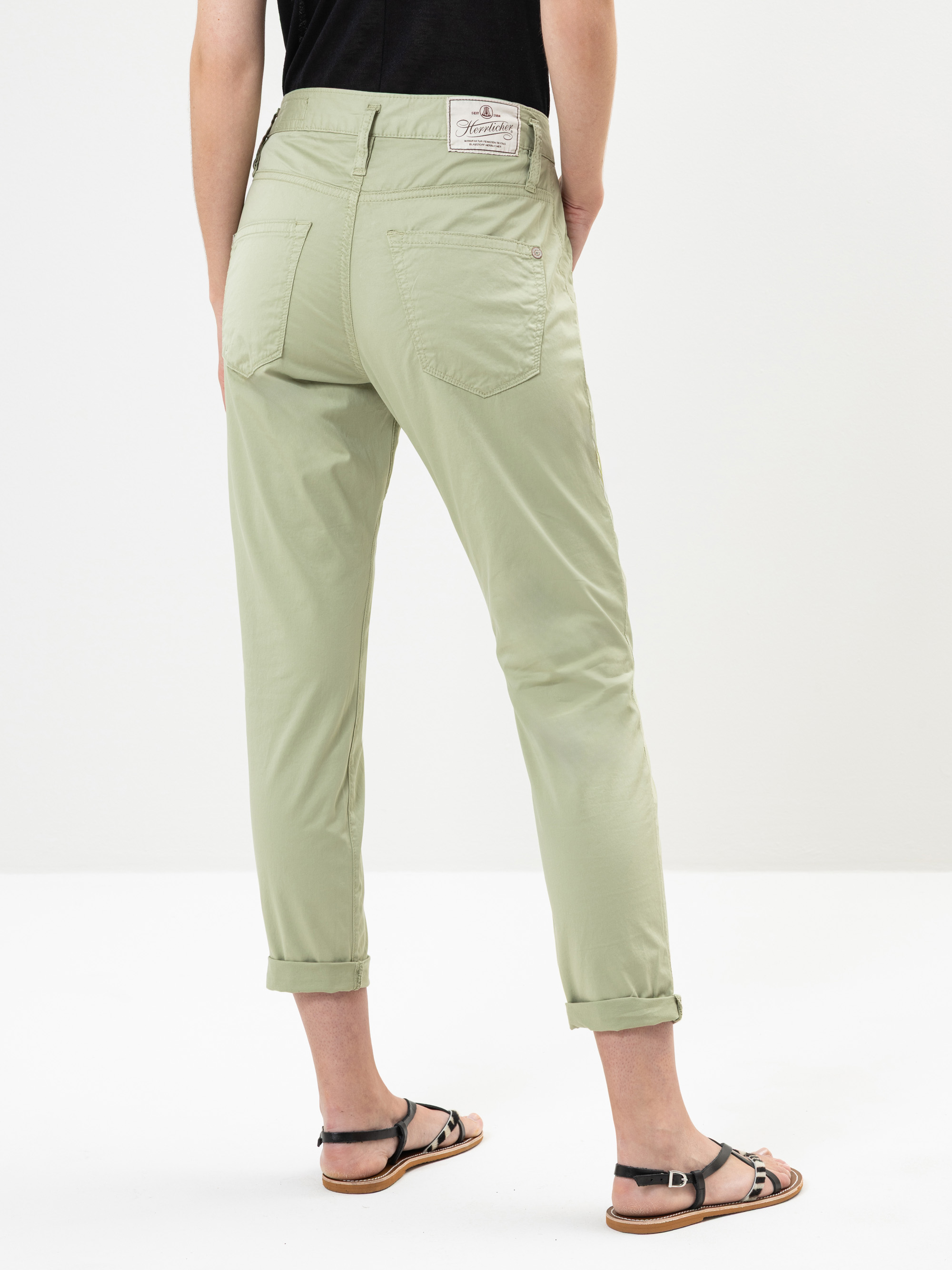 Eine Person, die eine hellgrüne Hose mit Bündchen, ein schwarzes Oberteil und schwarze Sandalen trägt, steht mit dem Rücken zur Kamera vor einem schlichten weißen Hintergrund.