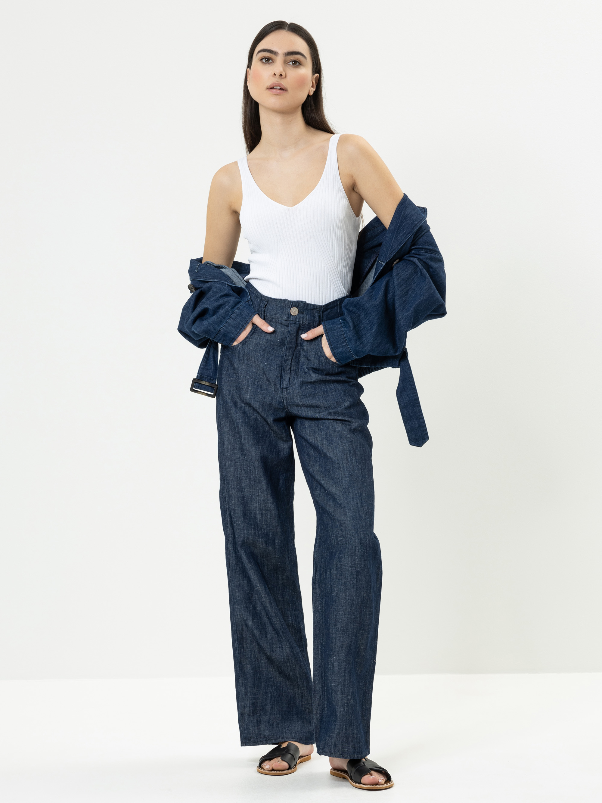 Eine Frau posiert vor einem schlichten weißen Hintergrund und trägt ein weißes ärmelloses Oberteil, Jeans mit weitem Bein, eine aufgeknöpfte Jeansjacke, die sie über die Arme gelegt hat, und schwarze Sandalen.