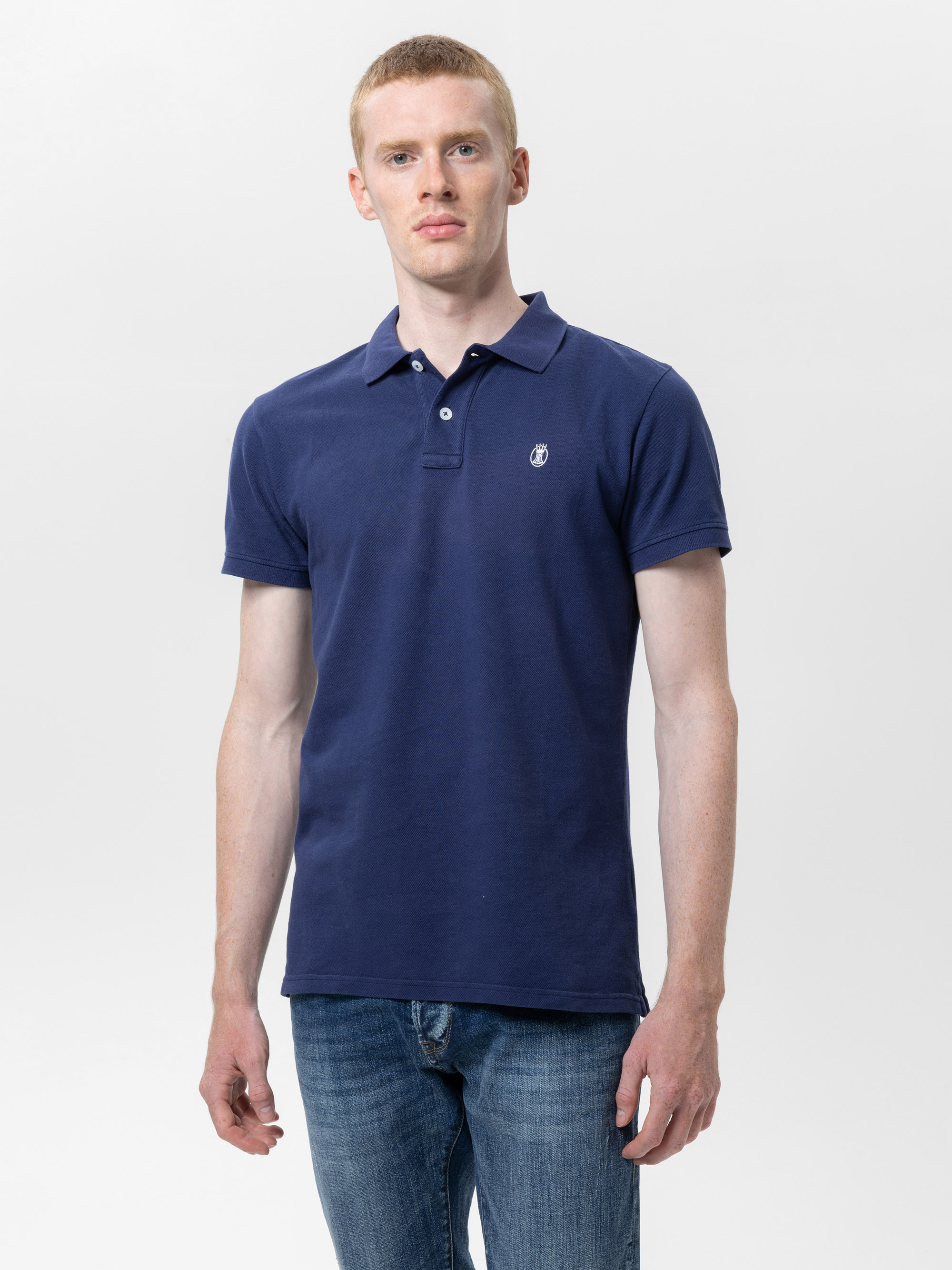 Ein Mann, der ein marineblaues Polohemd und blaue Jeans trägt, steht mit dem Gesicht nach vorne vor einem schlichten weißen Hintergrund.