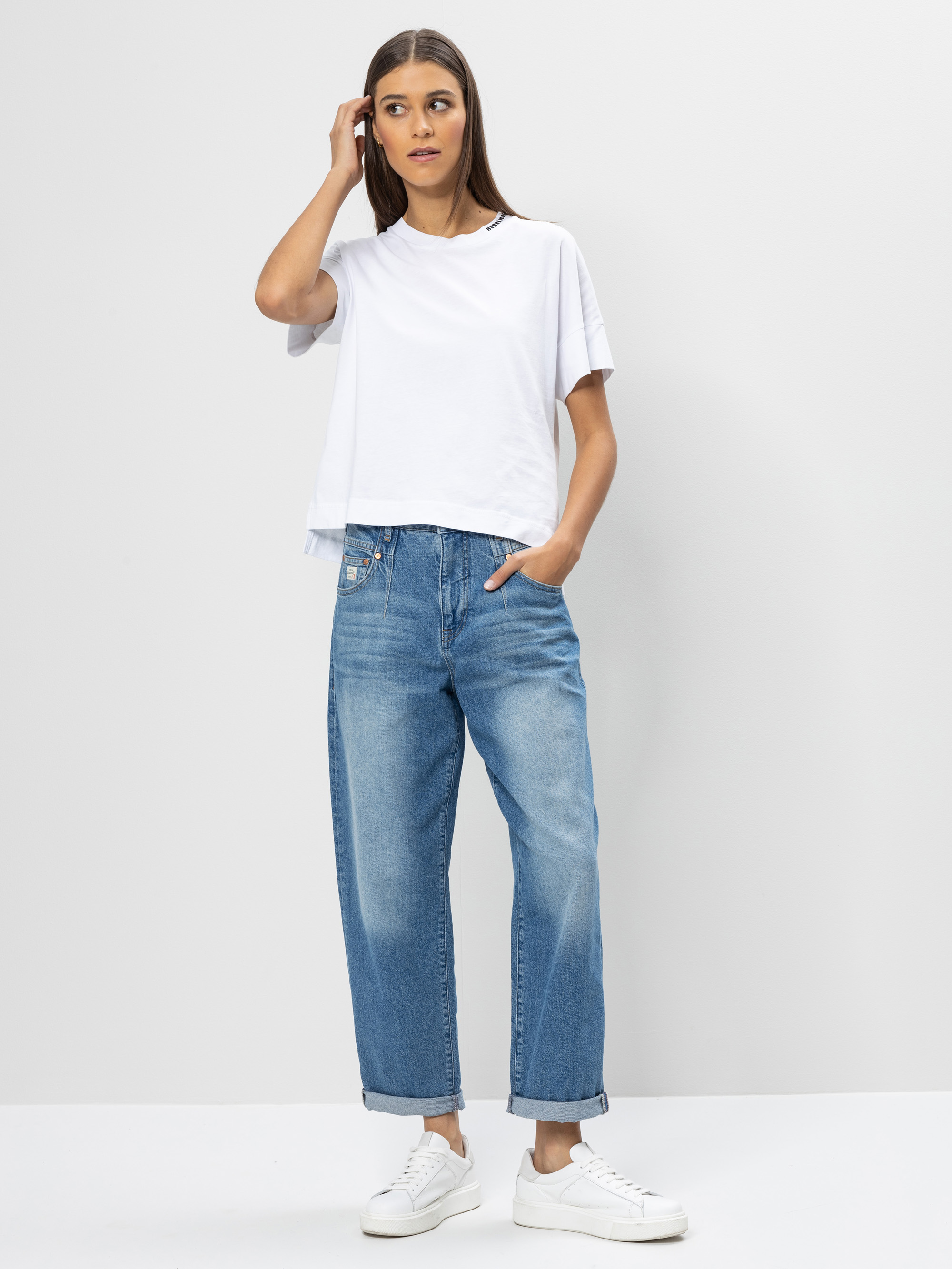 Eine Frau steht vor einem schlichten Hintergrund und trägt ein weißes T-Shirt, eine locker sitzende Bluejeans mit aufgerollten Bündchen und weiße Turnschuhe. Eine Hand steckt in ihrer Tasche, die andere berührt ihr Haar.