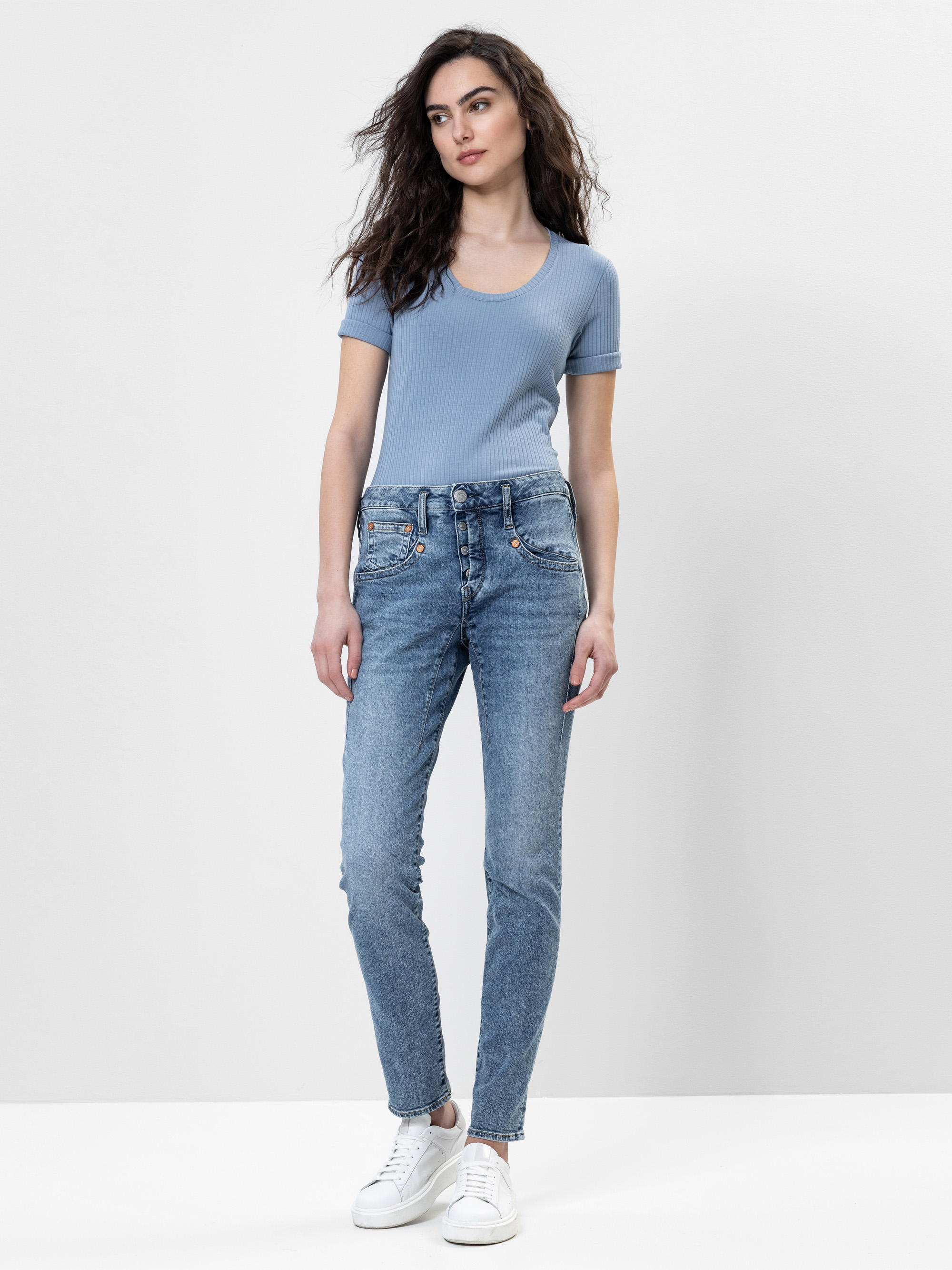 Eine Frau mit gewelltem dunklem Haar, die ein hellblaues kurzärmeliges Oberteil, blaue Jeans und weiße Turnschuhe trägt, steht vor einem schlichten weißen Hintergrund. Sie blickt nach vorne und hat die Hände in den Taschen.