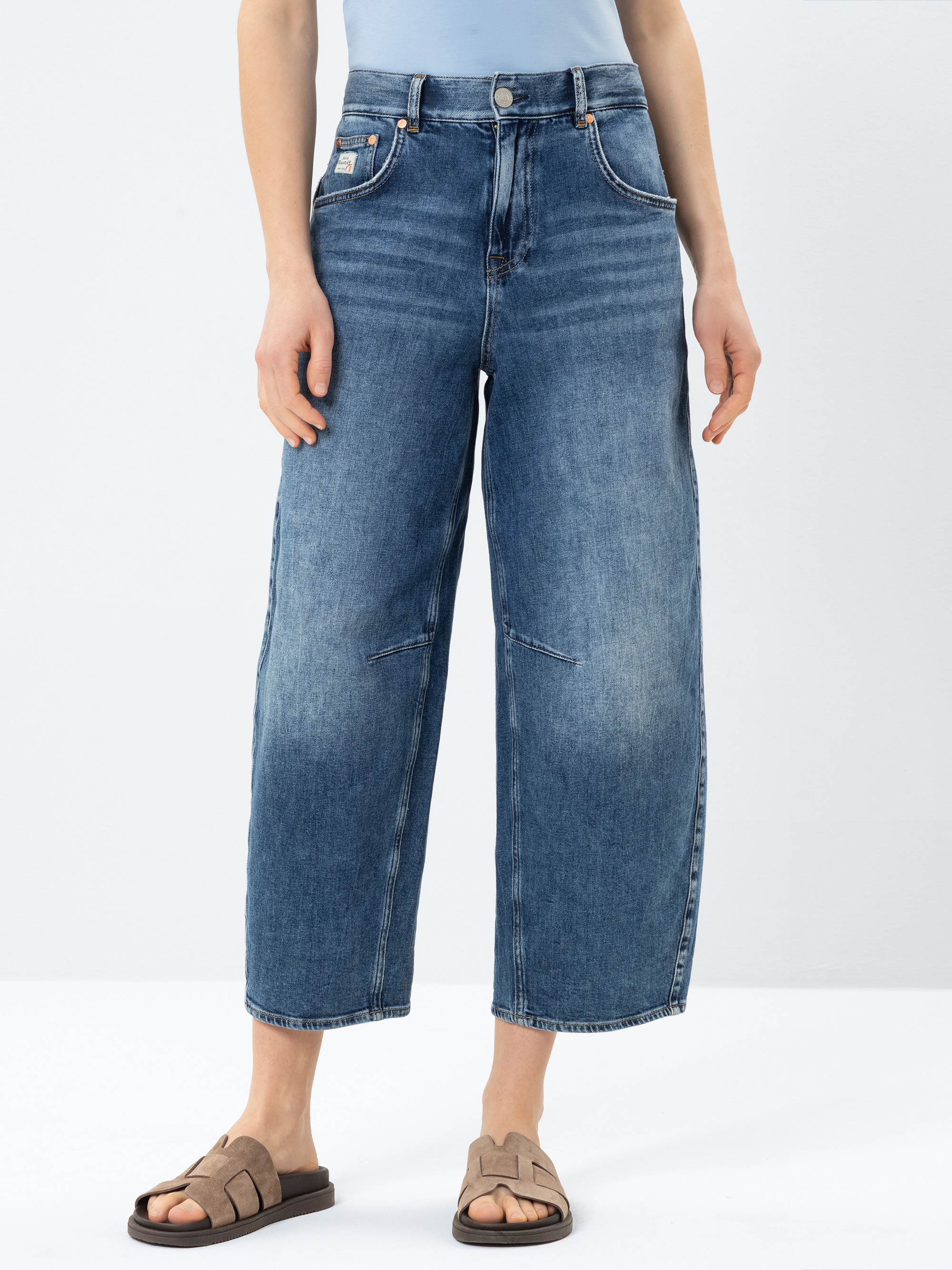 Stehende Person vor einfarbigem Hintergrund, bekleidet mit einer blauen Jeans mit hoher Taille und weitem Bein, einem hellblauen gesteckten Oberteil und beigen offenen Sandalen. Nur die untere Hälfte des Körpers ist sichtbar.