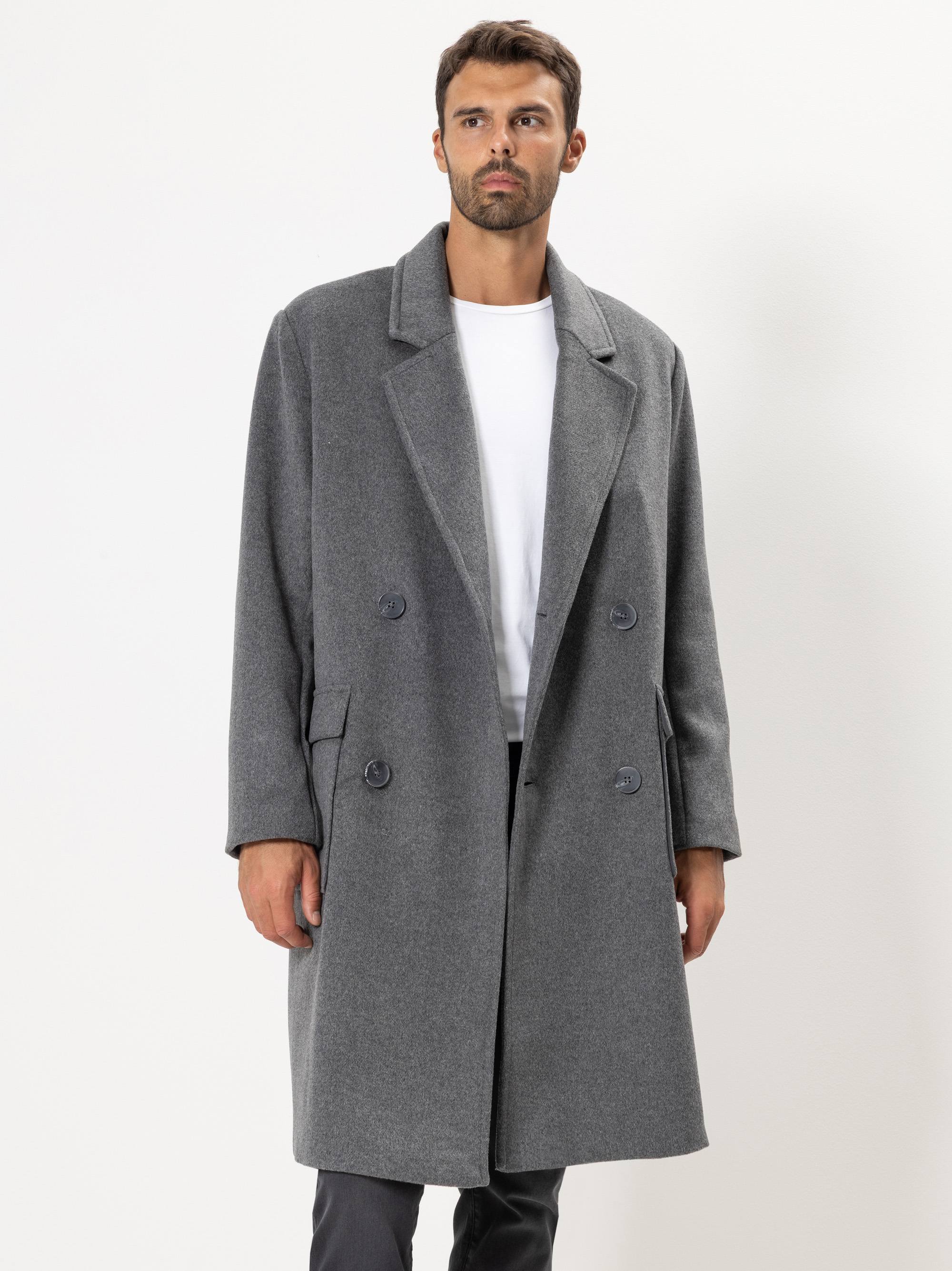 Ein Mann mit kurzen dunklen Haaren und Bart trägt einen langen grauen Zweireiher über einem weißen Hemd und einer dunklen Hose und steht vor einem schlichten weißen Hintergrund.